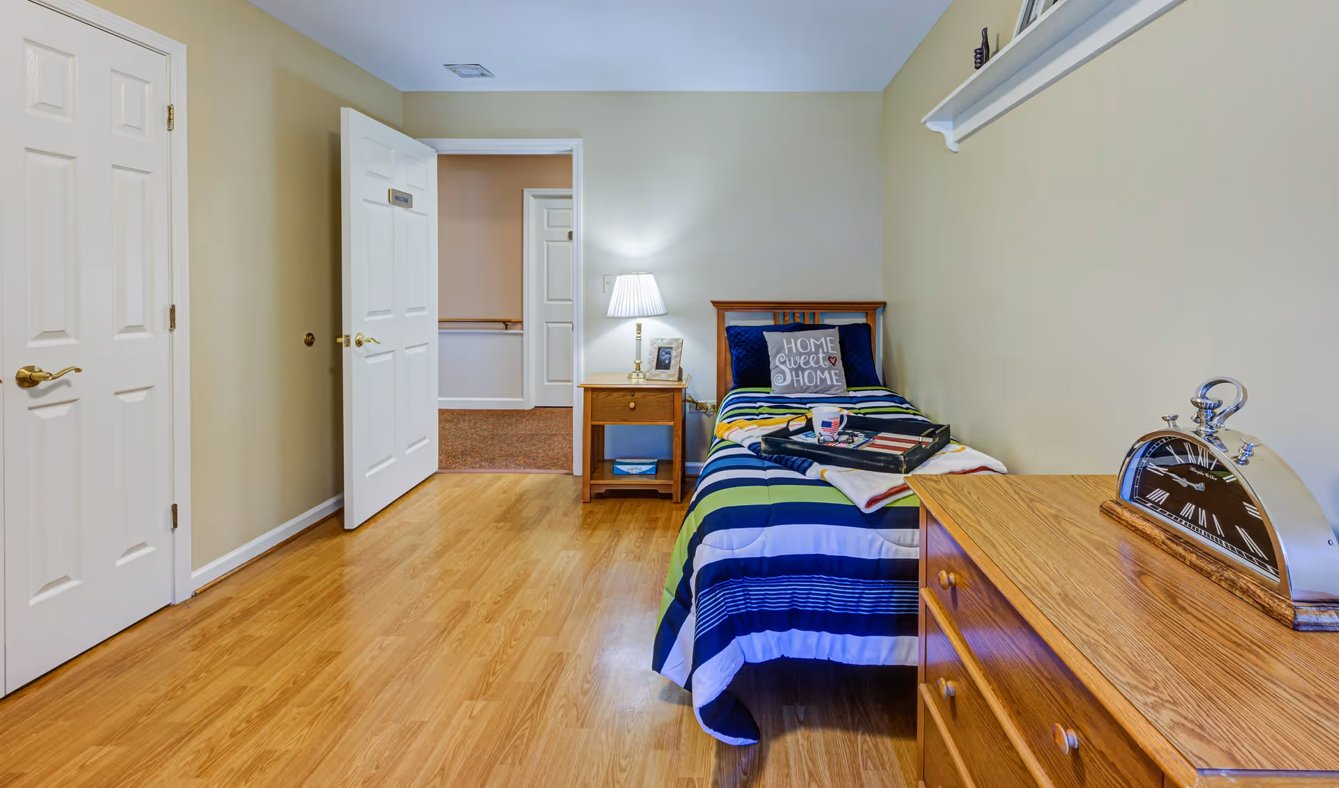 A small bedroom with a single bed covered in a striped blue, green, and white blanket. A pillow on the bed reads 'Home Sweet Home.' Next to the bed is a wooden nightstand with a lamp and a framed photo. Across from the bed is a wooden dresser with a silver clock on top. The room has light-colored walls and wooden flooring. Two white doors are visible, one open leading to a hallway.