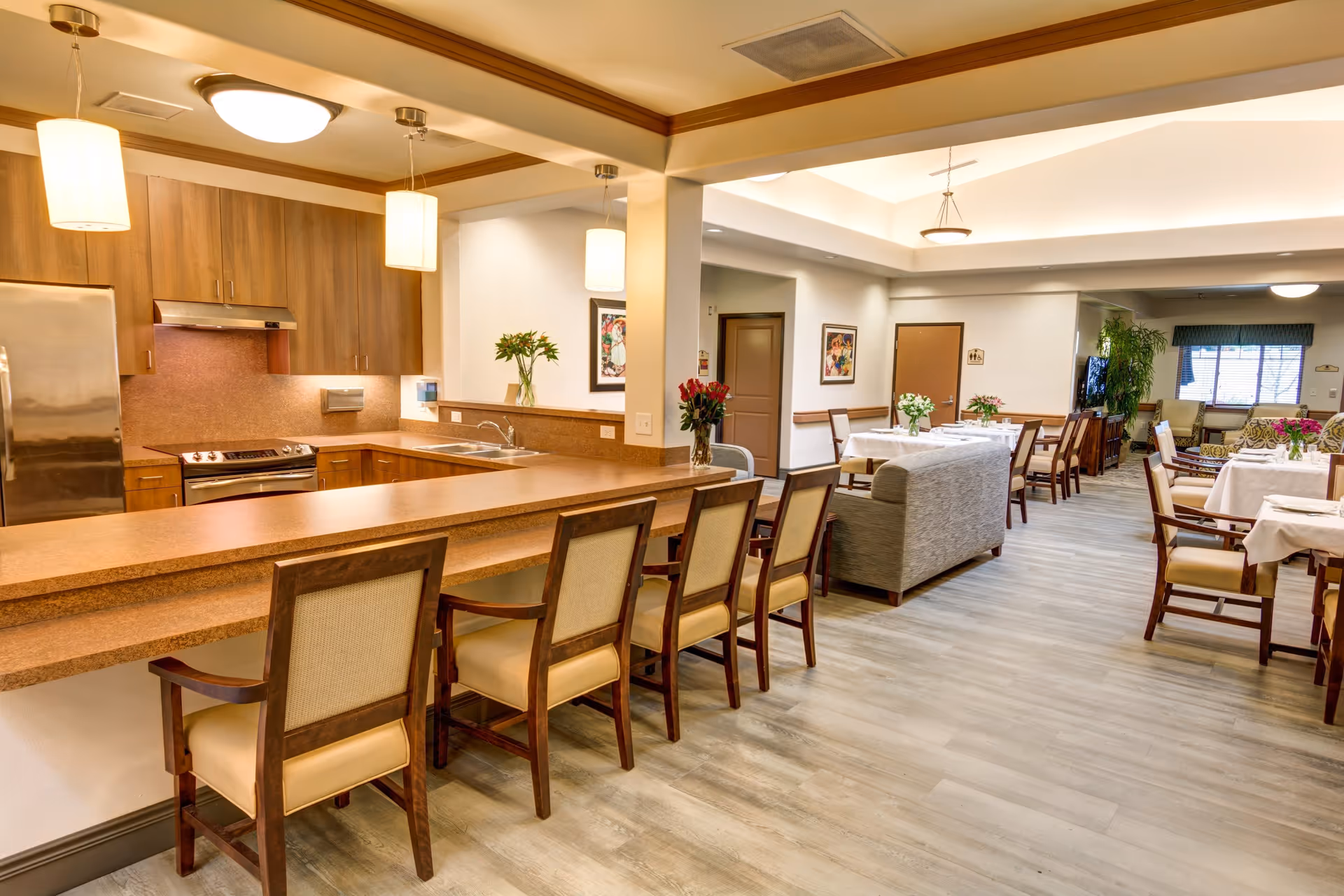 A spacious and well-lit common area in a senior living facility featuring a kitchen with wooden cabinets, a stainless steel refrigerator, and a stove. There is a long counter with several chairs for seating. Adjacent to the kitchen is a dining area with multiple tables covered with white tablecloths and chairs around them. A gray couch separates the kitchen and dining area. The room has light-colored flooring, ceiling lights, and decorative plants and flowers on the tables and counters.