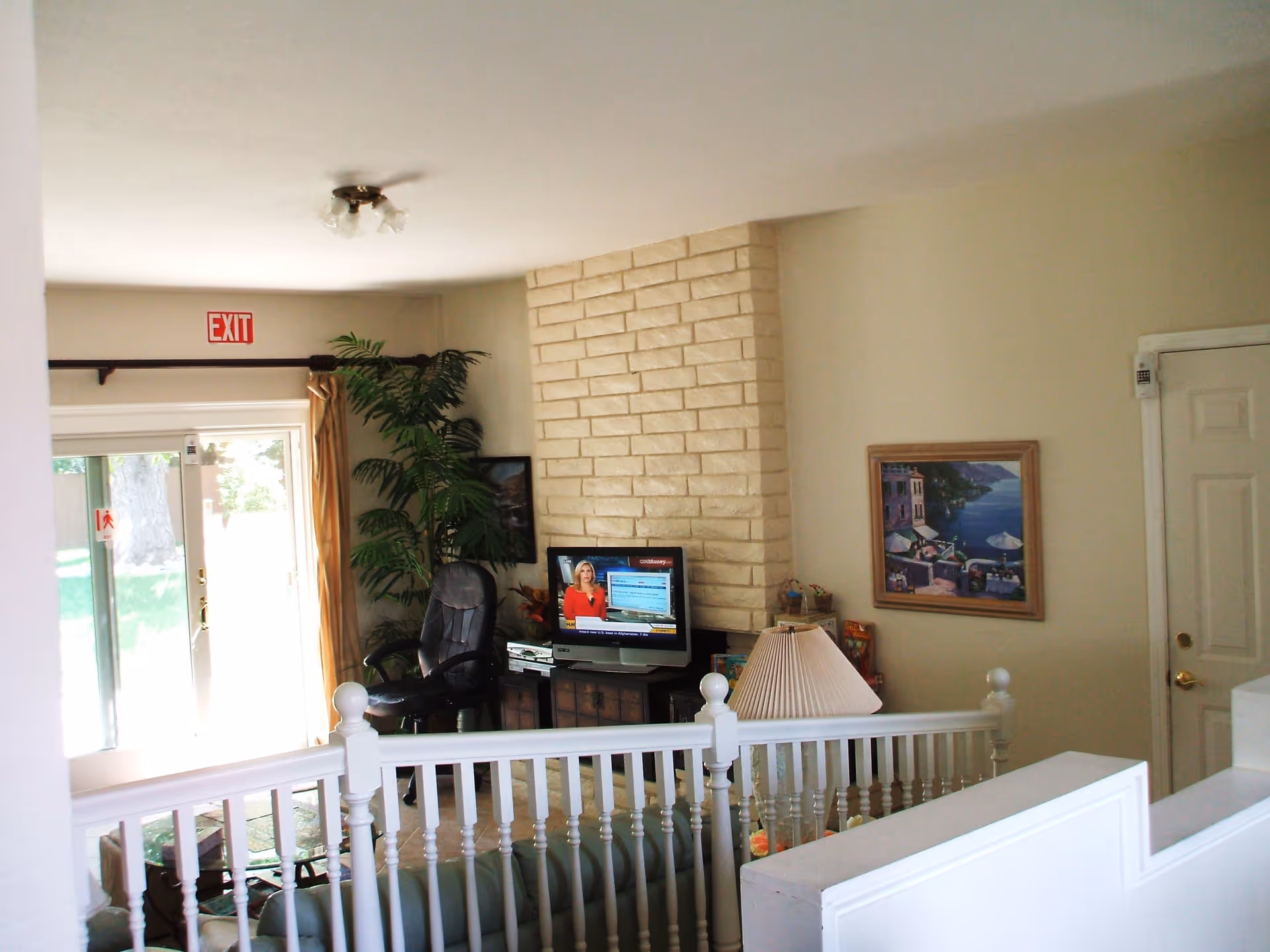 Interior view of a living room area with a white railing in the foreground, a stone fireplace with a TV on a stand showing a news channel, a black office chair, a large potted plant, a framed painting on the wall, a table lamp, and a glass sliding door with an exit sign above it.