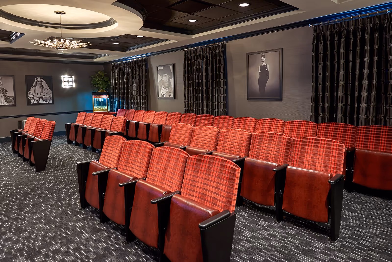 A small theater room with rows of red plaid upholstered seats arranged in front of a screen. The room has dark patterned curtains, black and white portraits on the walls, a popcorn machine in the corner, and a modern ceiling light fixture.