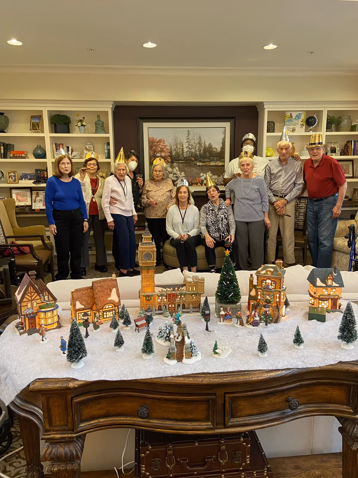 A group of elderly people wearing festive party hats standing and sitting in a living room with bookshelves and a large framed painting on the wall. In front of them is a wooden table decorated with a miniature winter village scene including small houses, trees, and figurines on a white cloth resembling snow.