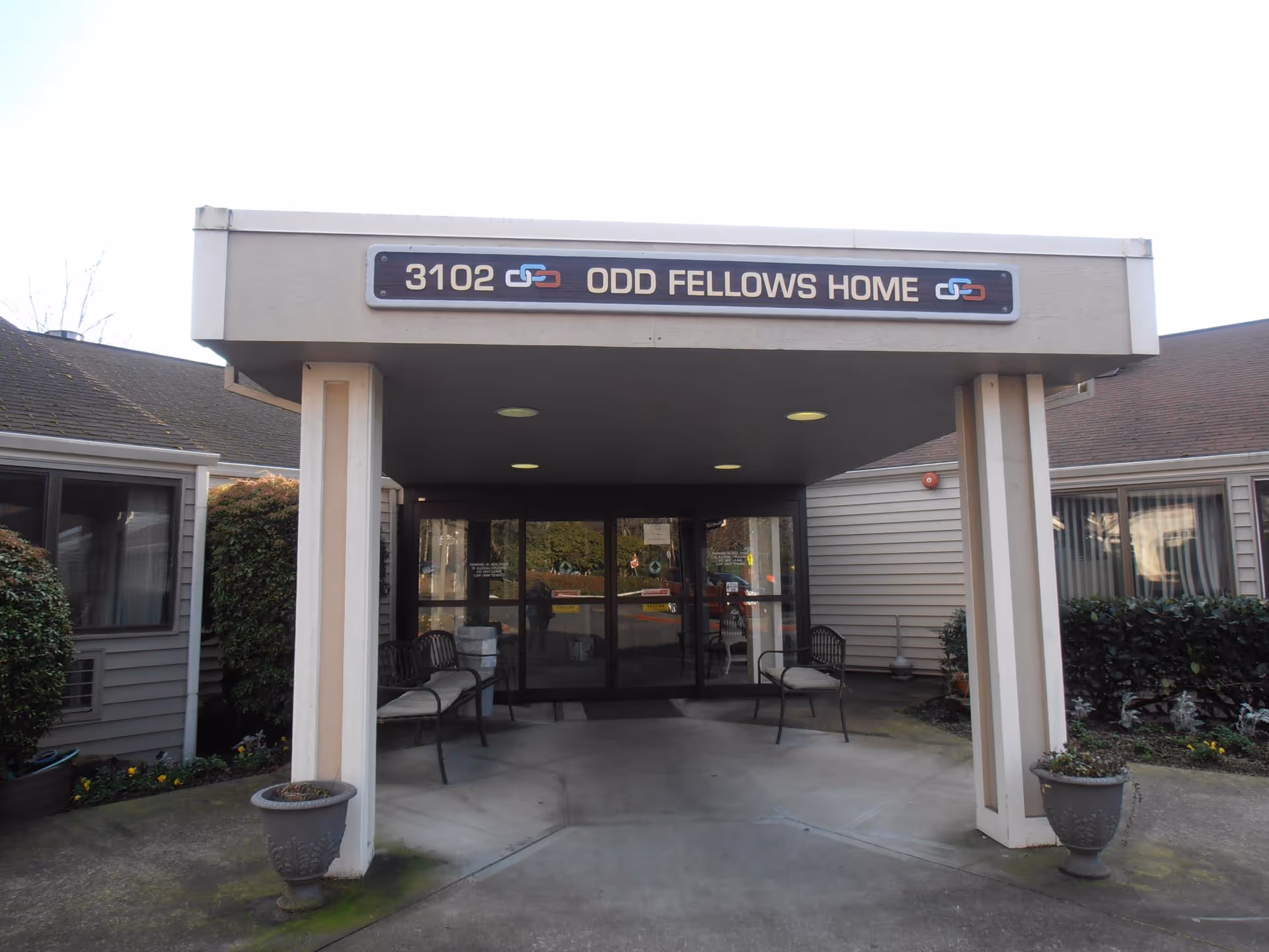Entrance to a building with a covered walkway supported by two columns. Above the entrance is a sign that reads '3102 ODD FELLOWS HOME'. There are benches on either side of the entrance and potted plants near the columns. The building has beige siding and windows with curtains.