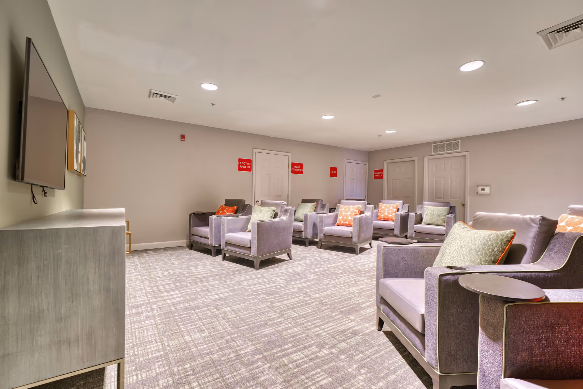 A spacious lounge area with multiple gray armchairs arranged in rows, each with decorative pillows in green and orange patterns. The room has a light gray carpet with a subtle pattern, beige walls, and several closed white doors with red signs above them indicating electrical panels and HVAC controls. A large flat-screen TV is mounted on the left wall above a gray cabinet.