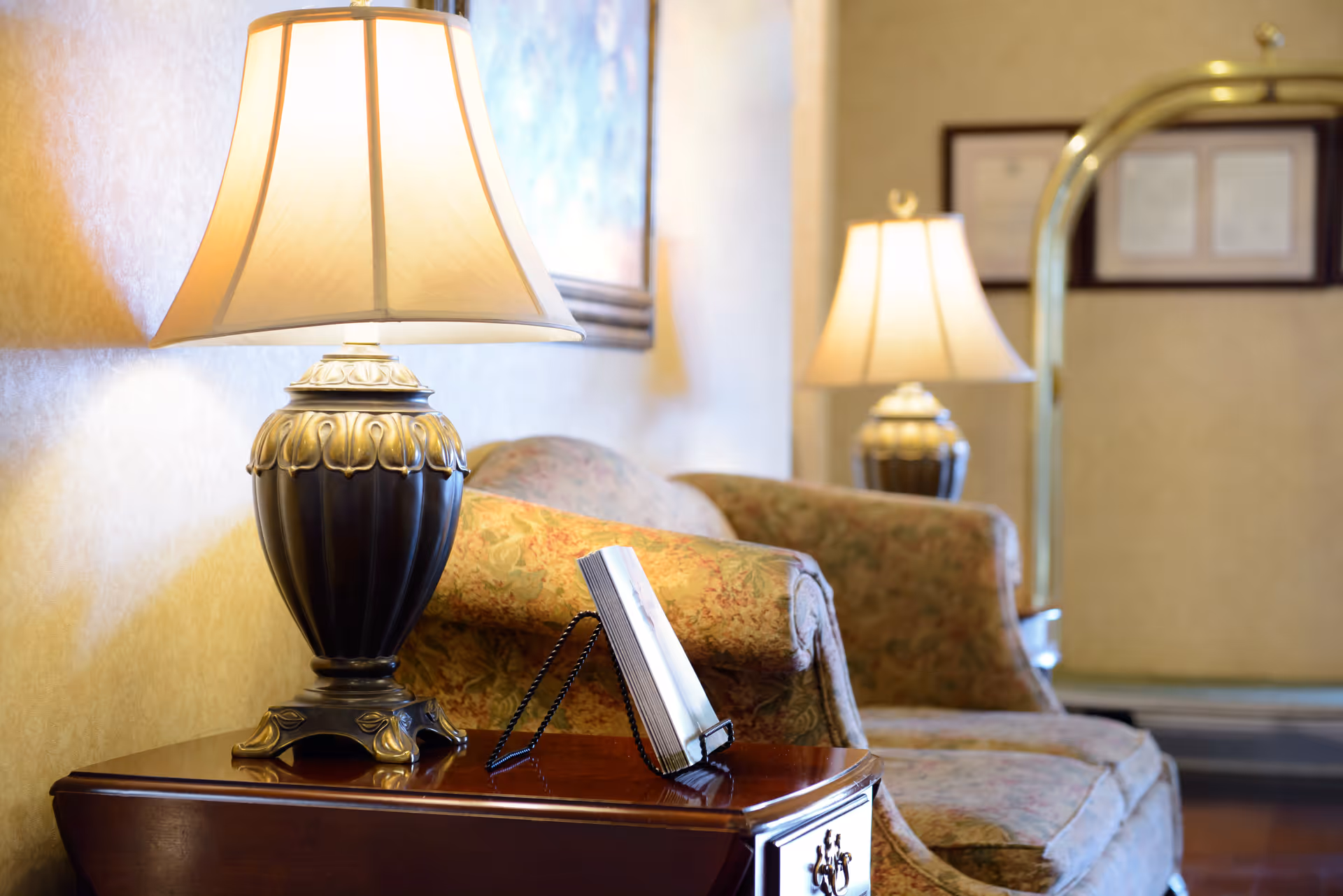 A cozy sitting area with a floral patterned sofa and two ornate table lamps on wooden side tables. There is a framed picture on the wall and a luggage cart in the background, suggesting a welcoming interior space.