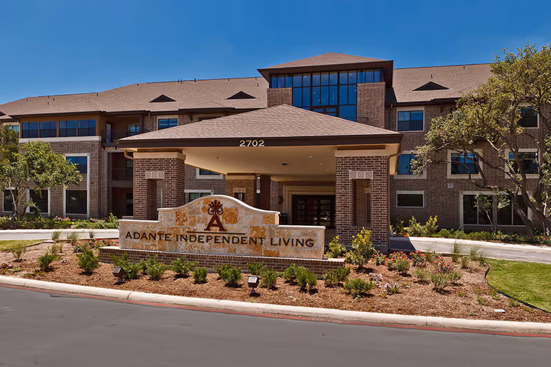 Front exterior view of Adante Independent Living facility showing a large brick building with multiple windows, a covered entrance with the address number 2702, and a landscaped area with shrubs and flowers in front of a stone sign displaying the facility name.