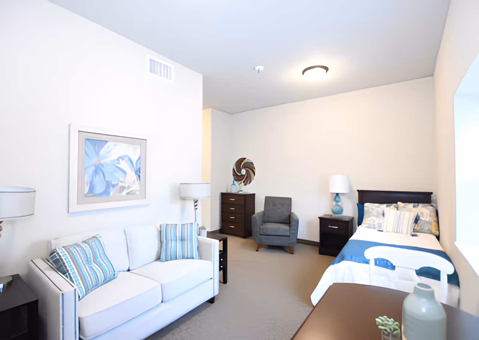 A bright and clean assisted living bedroom featuring a single bed with blue and white bedding, a white sofa with striped blue cushions, a gray armchair, a dark wood dresser and nightstand, two table lamps, and a framed artwork of blue flowers on the wall.