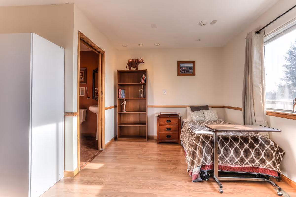 A small senior living bedroom with a single bed covered in a brown and white patterned blanket, a wooden nightstand with drawers, a wooden bookshelf with books and decorative items, and a window with beige curtains letting in natural light. There is a door leading to a bathroom with a visible sink and toilet. The room has light-colored walls and wooden flooring.