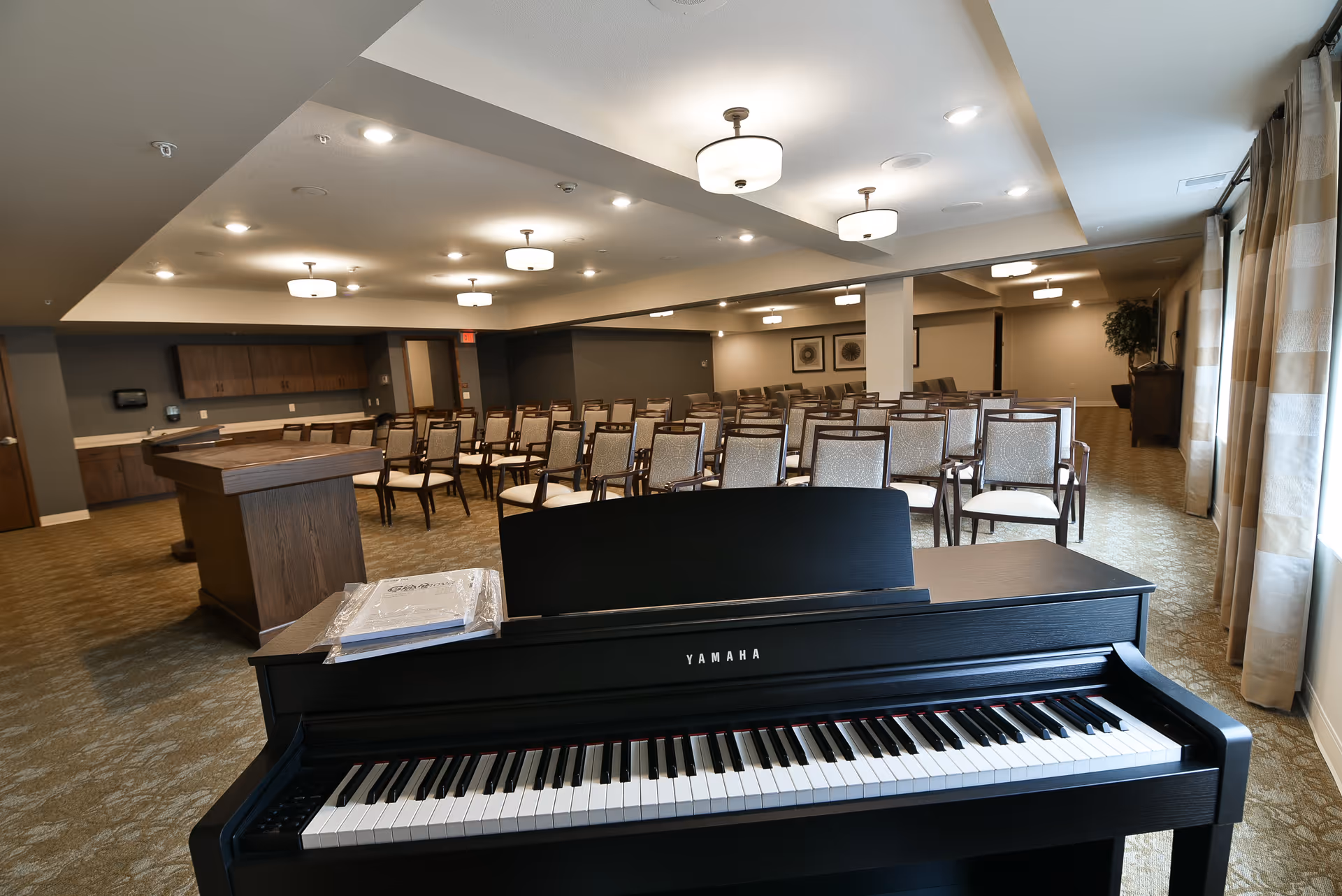 Large interior activity room with a Yamaha piano in the foreground and rows of chairs arranged for an audience.