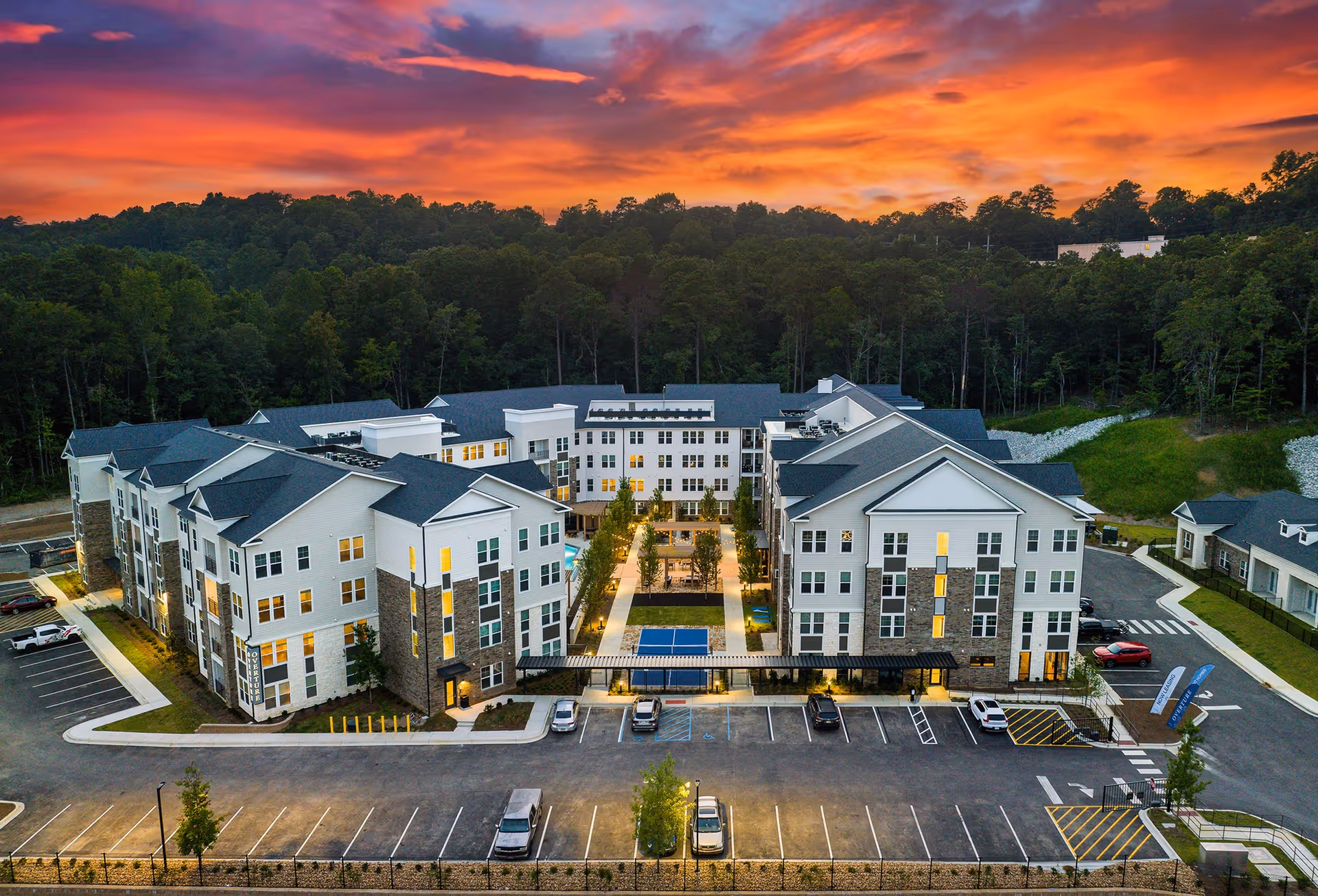 Aerial view of Overture Tributary senior living facility at sunset, showing a large, modern multi-story building with white and stone exterior, surrounded by trees and parking lots with several cars. The sky is vibrant with orange and purple hues.