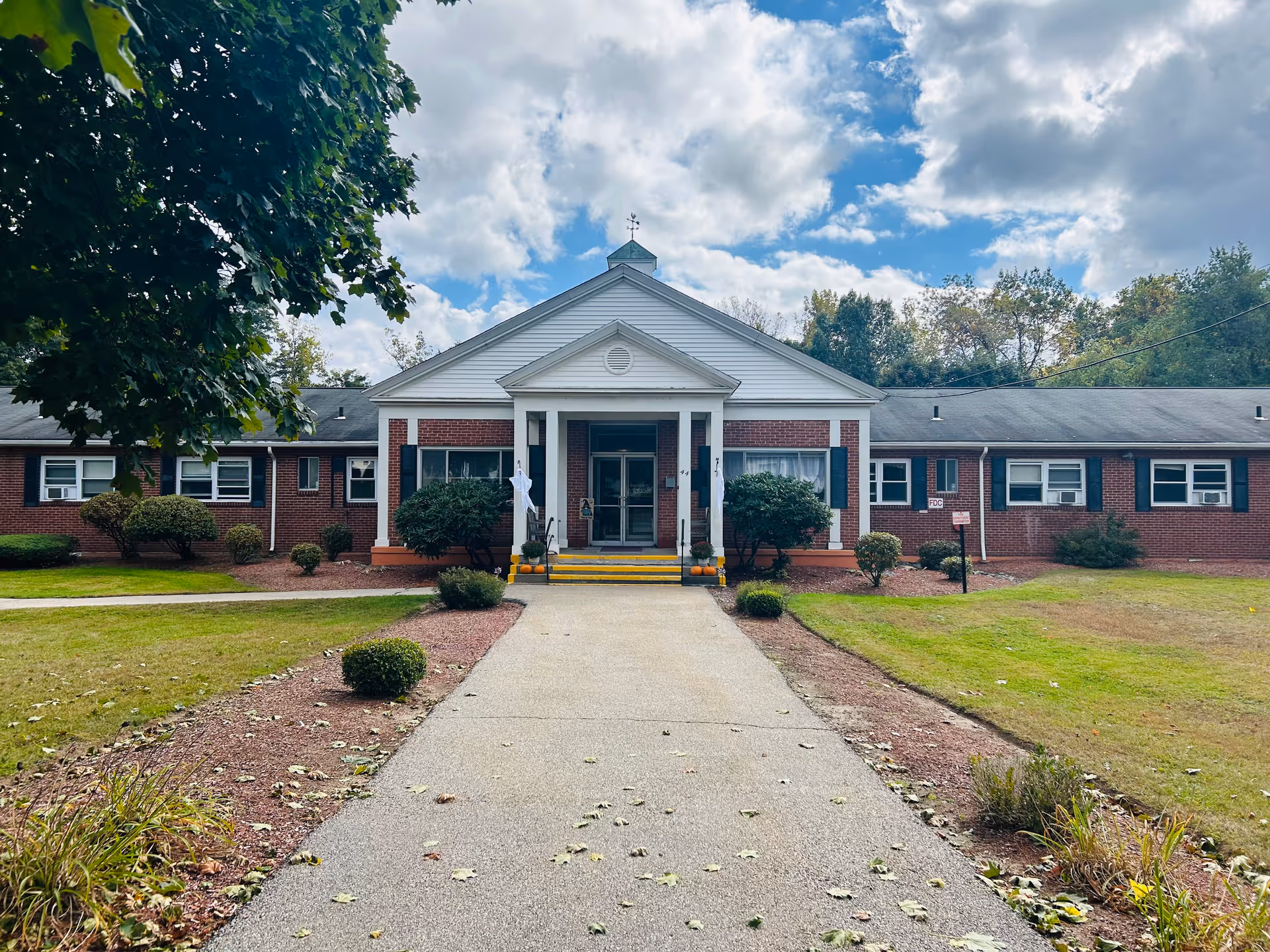 Front exterior view of a single-story brick building with white columns and a triangular pediment above the entrance. There is a paved walkway leading up to the entrance, surrounded by grass, bushes, and trees. The sky is partly cloudy with patches of blue.
