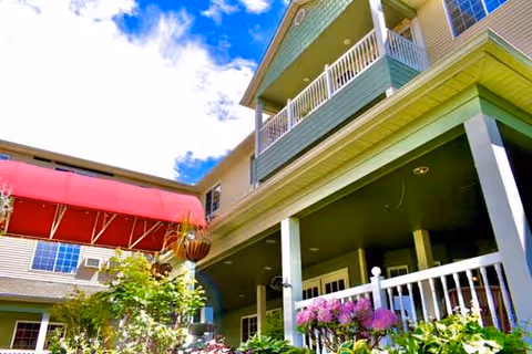Exterior view of a multi-story building with a covered porch and balcony, featuring white railings and surrounded by greenery and flowers under a partly cloudy blue sky.