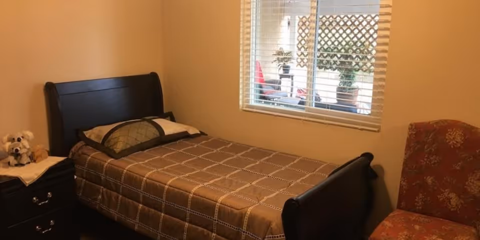 A small bedroom with a single bed covered in a brown checkered bedspread, a pillow, a nightstand with two stuffed animals, a window with blinds showing an outdoor patio with plants, and an upholstered chair with a floral pattern.