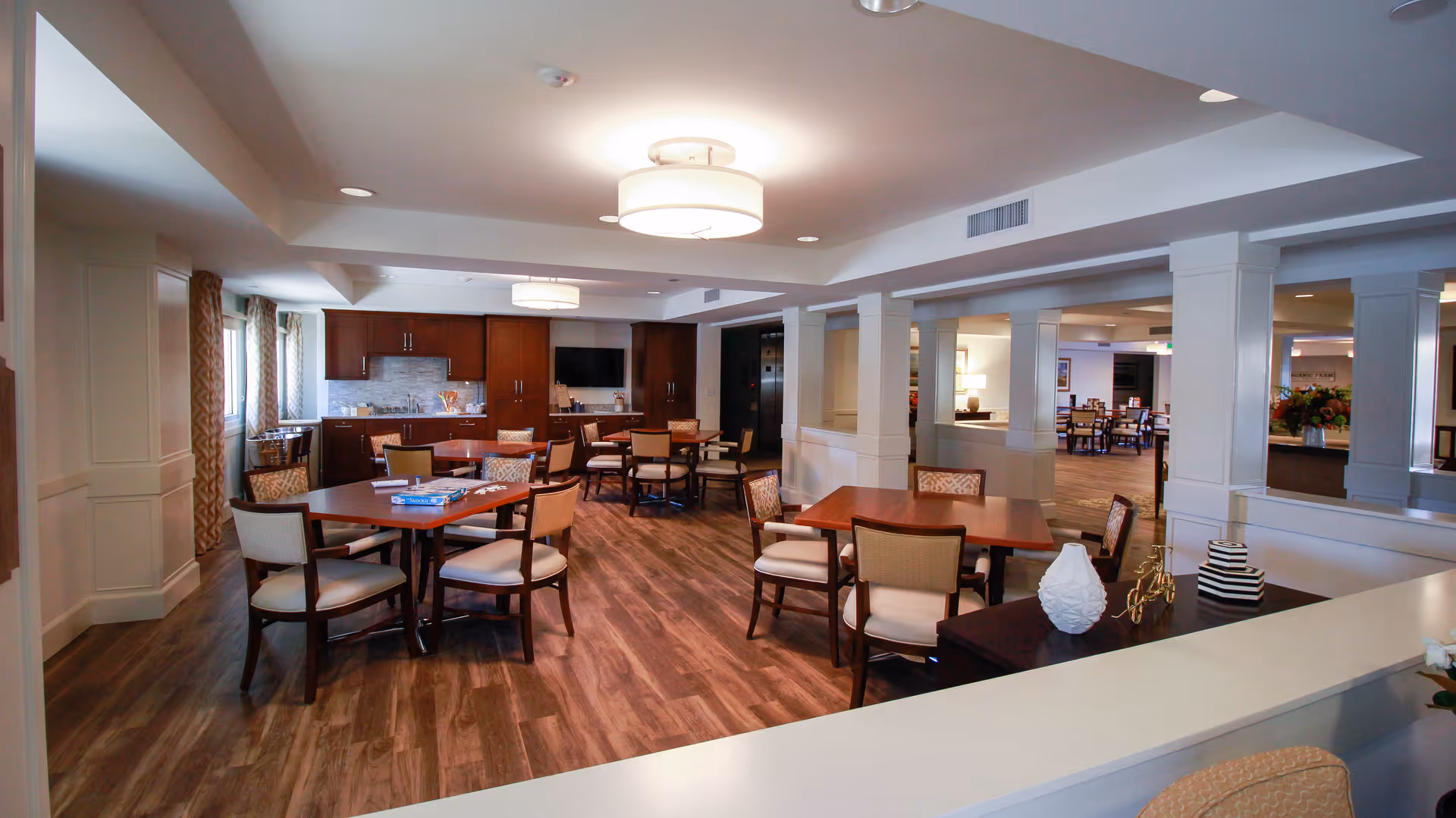 A spacious dining area in a senior living facility with multiple wooden tables and cushioned chairs arranged neatly. The room features wood flooring, modern ceiling lights, and a kitchenette with cabinets and a countertop in the background. Large windows with patterned curtains allow natural light to enter the room.