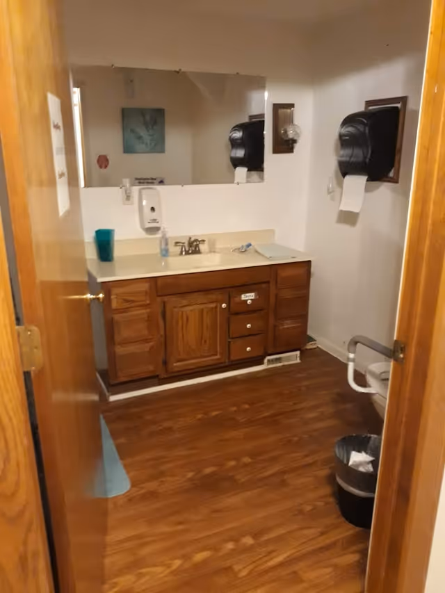 View into a bathroom with wooden floor and wooden vanity with a sink. There is a large mirror above the sink, a soap dispenser on the wall, a blue cup on the counter, and a paper towel dispenser mounted on the wall. A trash can is visible on the floor next to a toilet with a grab bar.