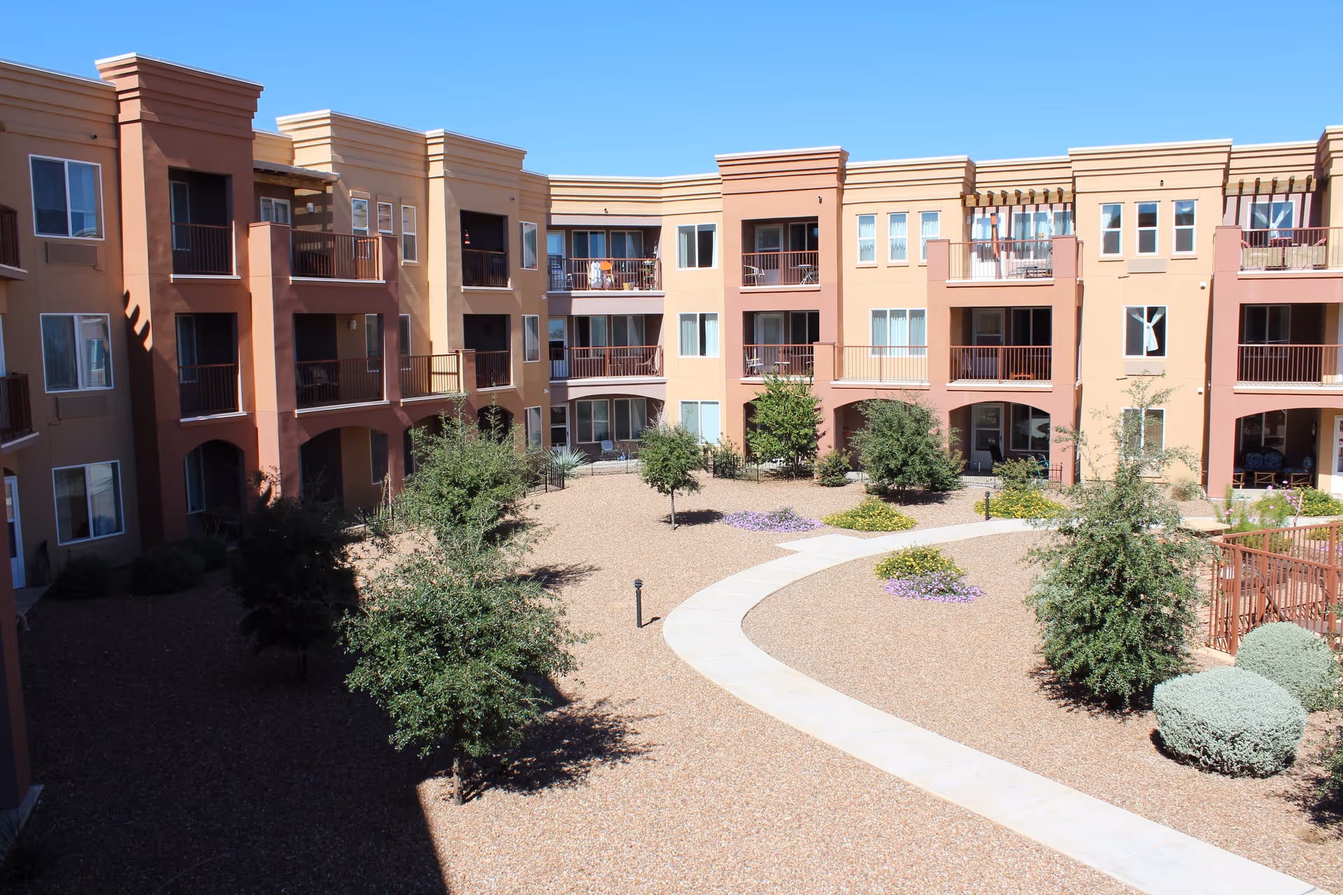 View of a multi-story residential building surrounding a landscaped courtyard with small trees, bushes, and a curved concrete walkway under a clear blue sky.
