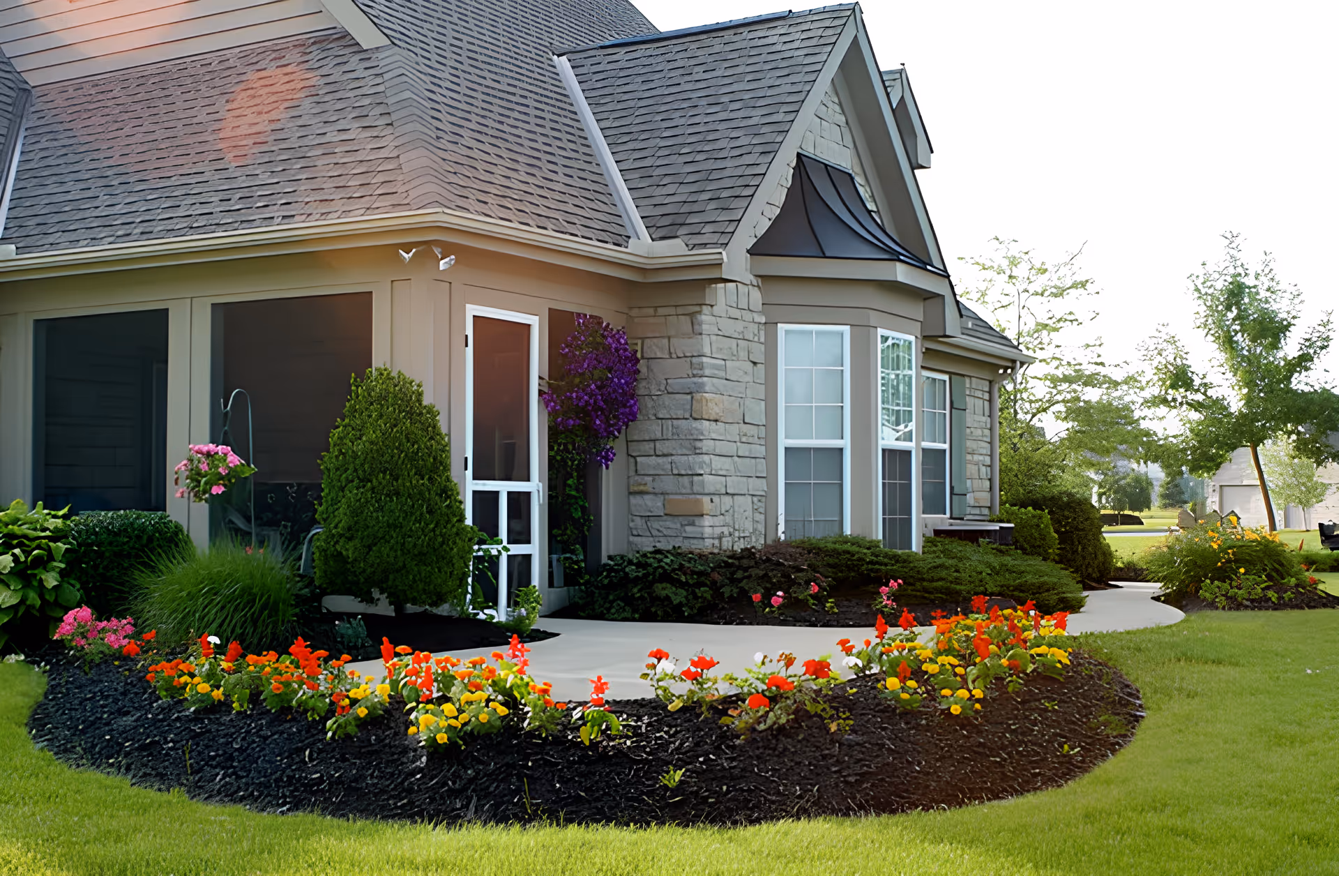 Exterior view of a residential-style building with a stone and siding facade, a shingled roof, and large windows. The entrance features a screened door surrounded by well-maintained landscaping including colorful flowers, bushes, and green grass. Trees and additional greenery are visible in the background.