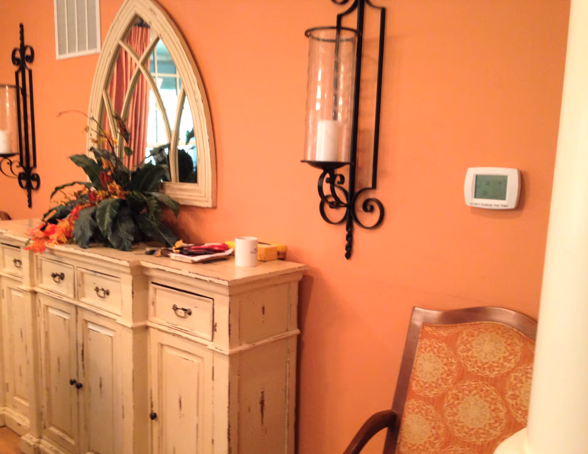 Interior view of a room with peach-colored walls featuring a distressed white wooden sideboard with drawers and cabinets. On top of the sideboard is a decorative arrangement of green and orange foliage, a white mug, and some small items. Above the sideboard is an arched mirror with a white frame. A black wrought iron candle holder with a glass hurricane and white candle is mounted on the wall. To the right, there is a thermostat and a wooden chair with an orange patterned cushion.