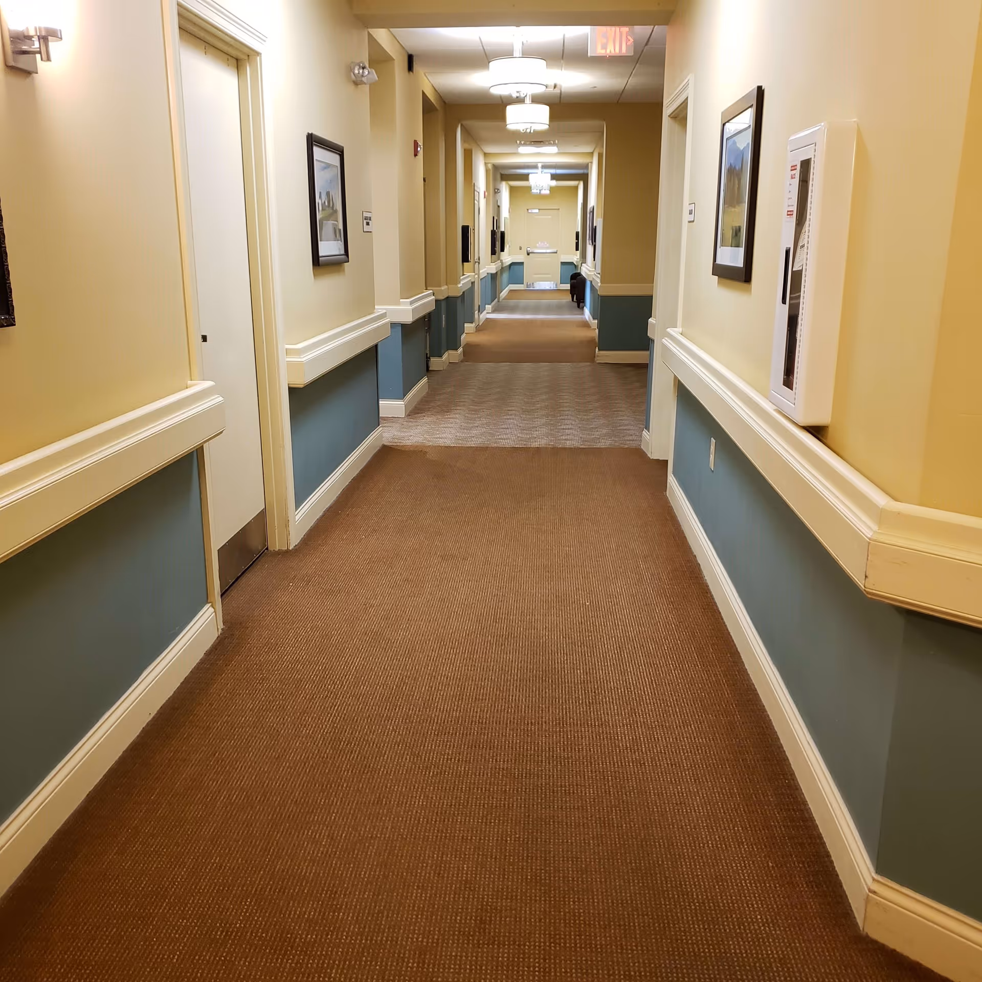 A long, well-lit hallway in a senior living facility with beige and blue walls, brown carpet, framed pictures on the walls, and ceiling lights. Doors line both sides of the hallway, and an exit sign is visible at the far end.