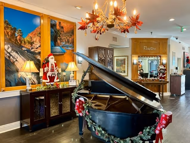 Interior view of a senior living facility lobby decorated for the holidays with a black grand piano adorned with garlands and red stockings. There are two table lamps on a cabinet with a Santa Claus figurine, a large scenic wall mural, a chandelier with red leaves, and a reception desk in the background with holiday decorations.