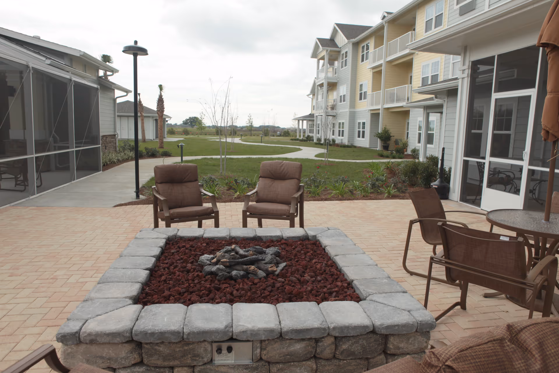 Outdoor patio area at The Carriage House Gracious Retirement Living featuring a square stone fire pit surrounded by cushioned chairs and a round table with chairs. The patio is paved with bricks and adjacent to a multi-story residential building with balconies and screened porch areas. There is a grassy area with a walking path and some young trees in the background under a cloudy sky.