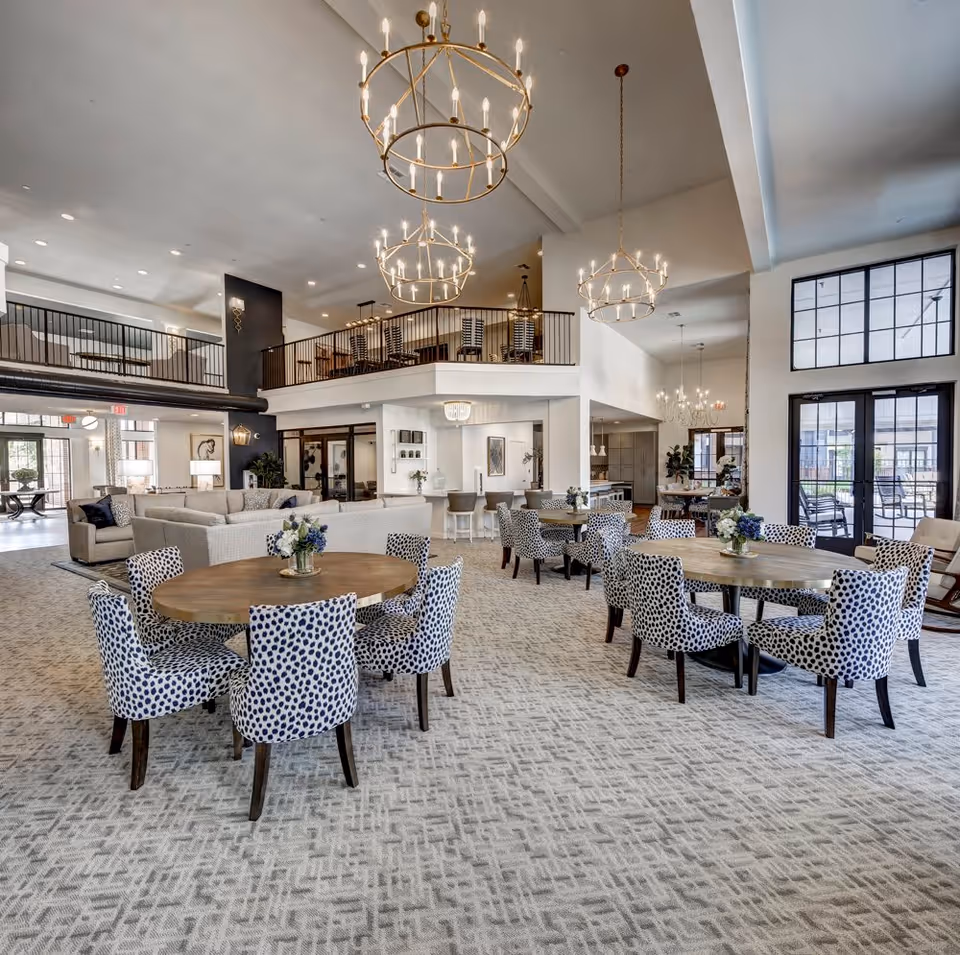 Spacious and well-lit common area in a senior living facility featuring multiple round wooden tables surrounded by blue and white patterned chairs. The room has high ceilings with large, circular chandeliers, a carpeted floor with a geometric pattern, and large windows and glass doors allowing natural light to enter. There is a seating area with sofas in the background and a mezzanine level with additional seating.