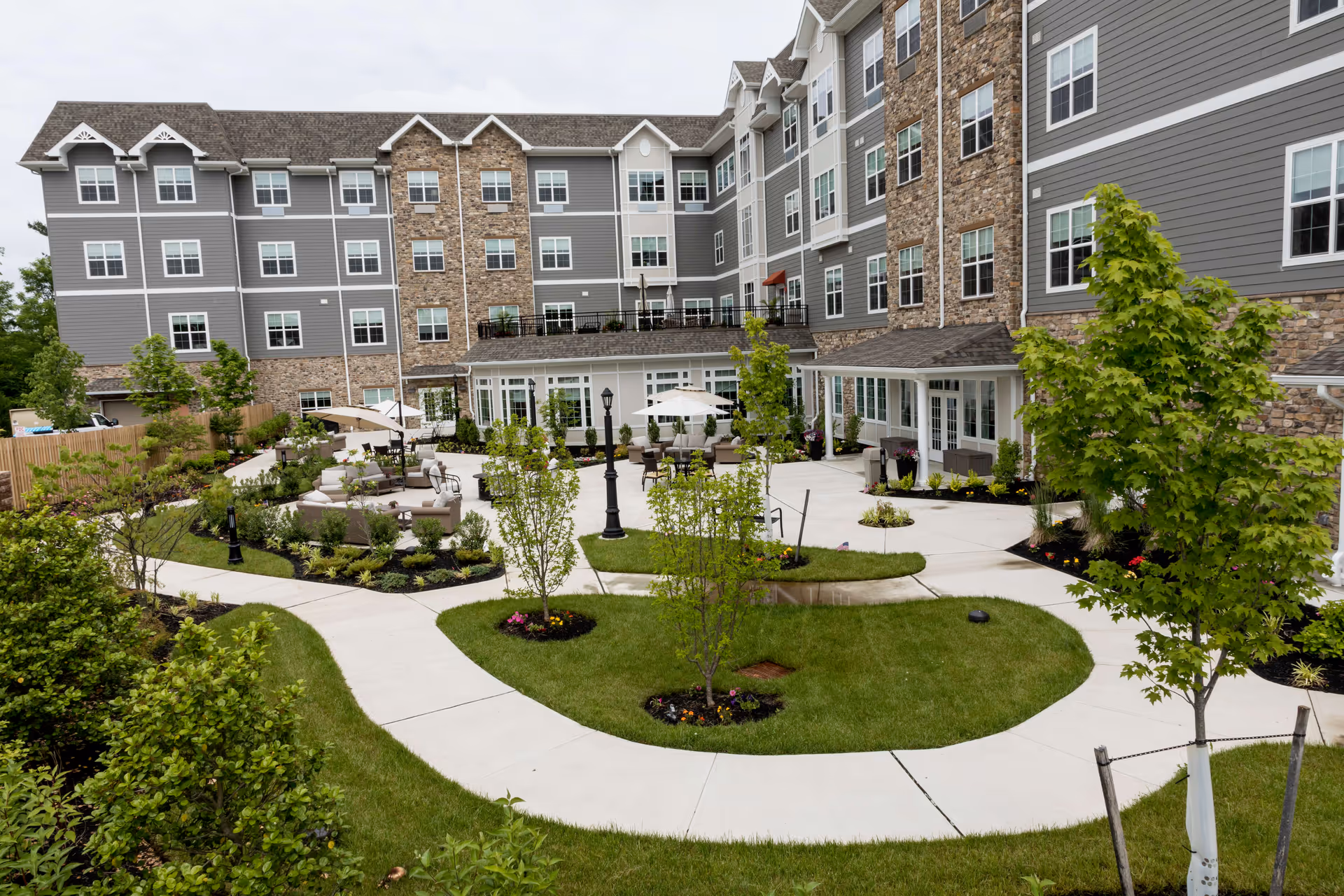 Landscaped courtyard with winding sidewalks, seating areas and young trees in front of a multi-story senior living building.