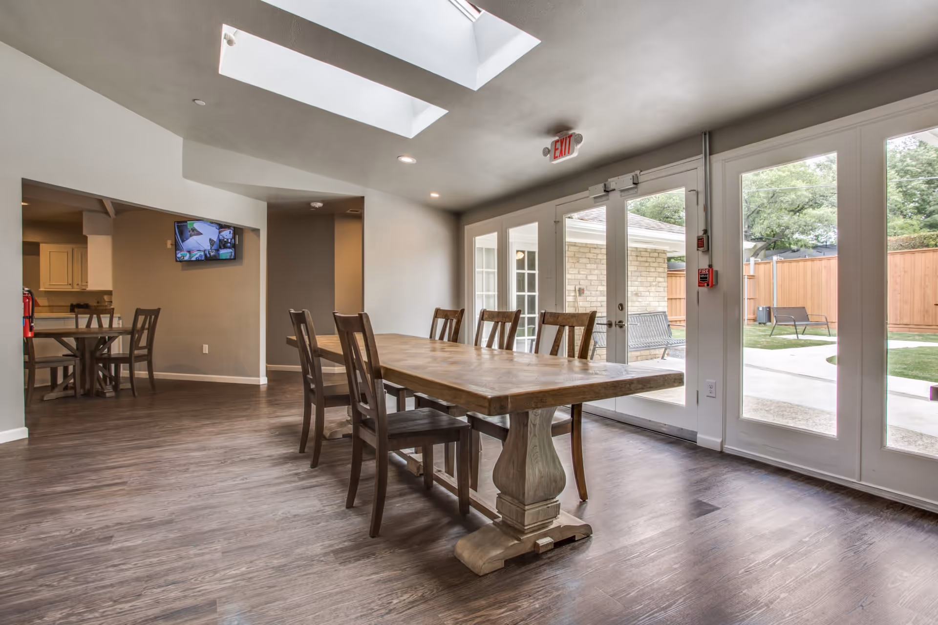 A bright dining area with a large wooden table and six chairs on a wood floor. The room has skylights in the ceiling and large glass doors leading to an outdoor patio with benches and a wooden fence. A smaller table with chairs and a wall-mounted TV are visible in the background.