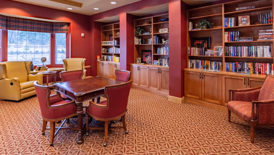 A cozy reading and sitting area in a senior living facility with red walls and carpet. There is a wooden table surrounded by four red upholstered chairs in the center. To the left, two yellow armchairs are positioned near a window with plaid valances. The back wall features built-in wooden bookshelves filled with books and decorative items.