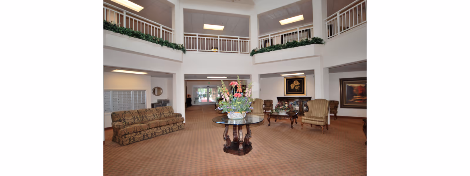 Spacious two-story lobby with seating, a central table topped with a floral arrangement, artwork on the walls, and upper balcony railings.