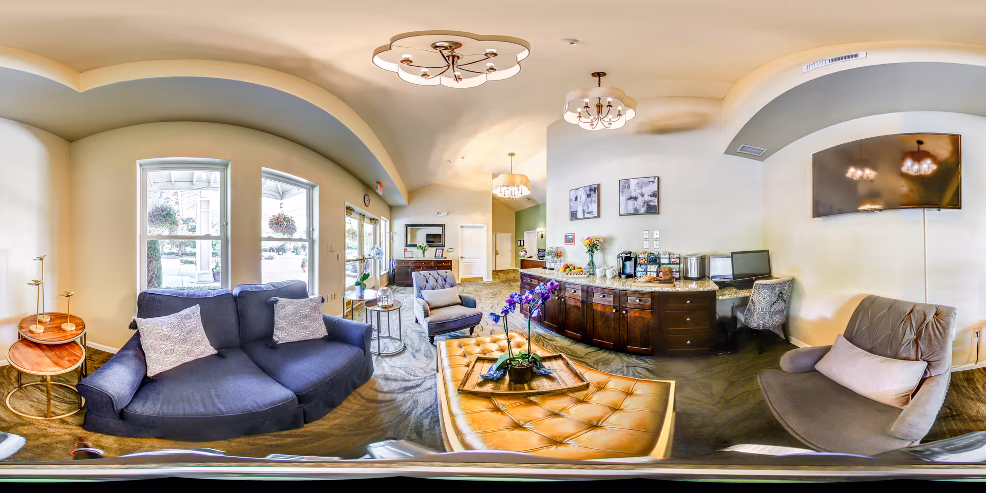A bright and spacious living room area in a senior living facility with large windows letting in natural light. The room features a blue sofa with two patterned pillows, a gray armchair with a light cushion, a tufted leather ottoman with a decorative tray and purple orchid, and a wooden side table. The back wall has a countertop with coffee and snack stations, a computer workstation, and two abstract paintings. A flat-screen TV is mounted on the wall above the armchair. The ceiling has multiple modern light fixtures and arched architectural details.