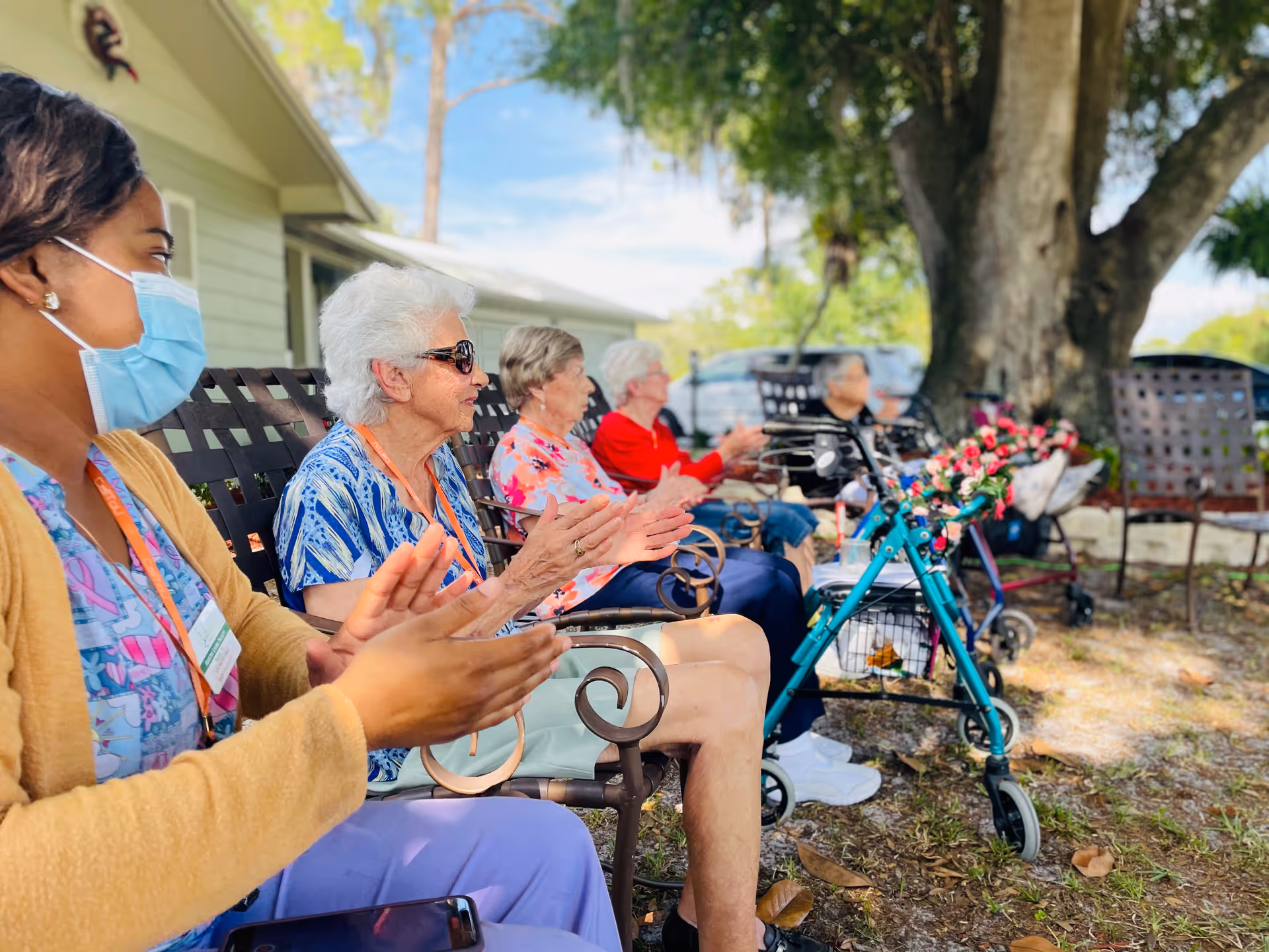 A group of elderly women and a caregiver sitting outdoors on metal chairs, clapping and enjoying an activity. The caregiver is wearing a face mask and a yellow cardigan. There are trees and a building in the background, and a walker decorated with flowers is visible in the foreground.