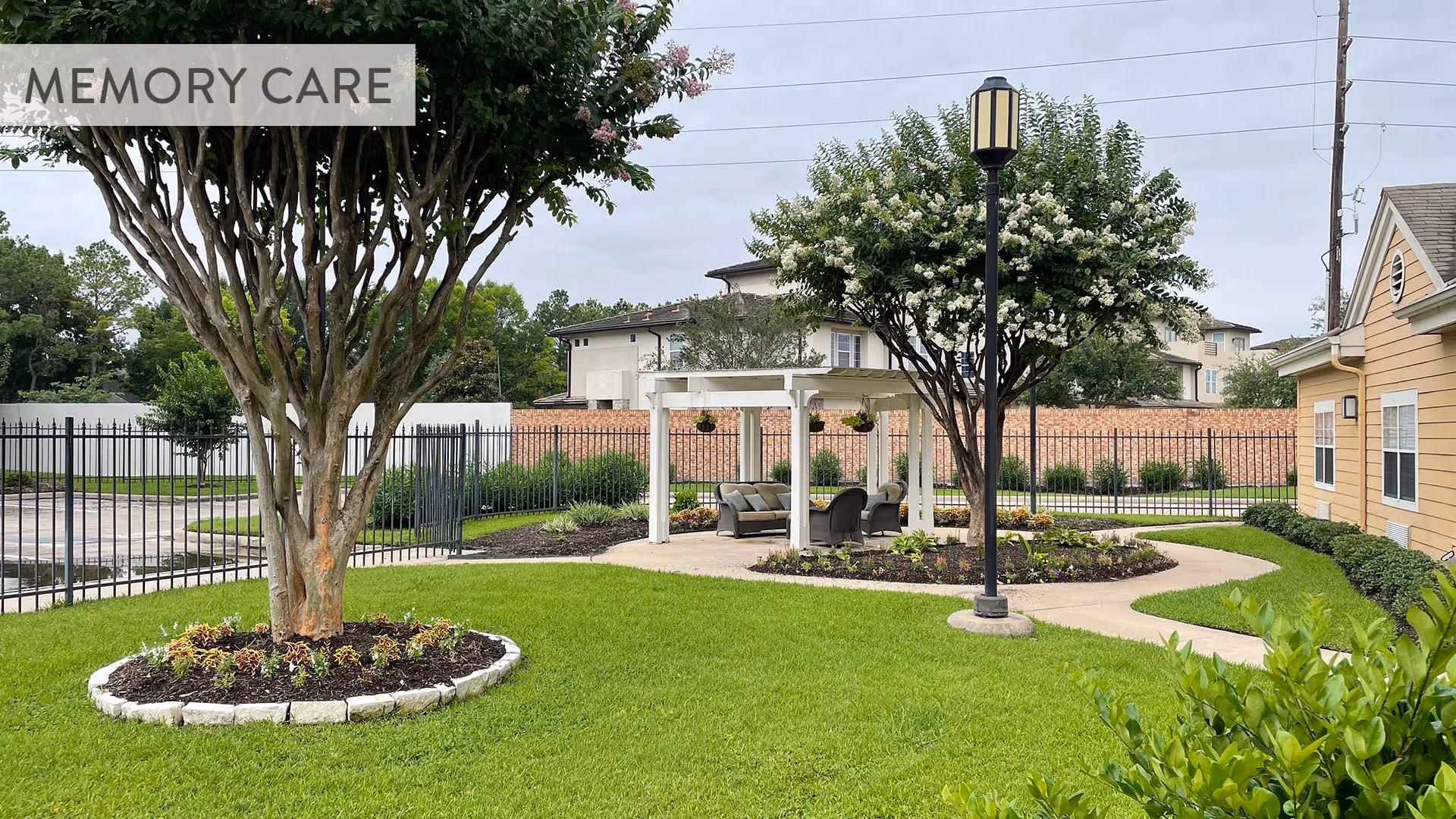 Outdoor garden area with green grass, two trees with flower beds, a black lamp post, and a white pergola with cushioned chairs underneath. A black metal fence and residential buildings are visible in the background. The image has a semi-transparent overlay in the top left corner with the text 'MEMORY CARE'.