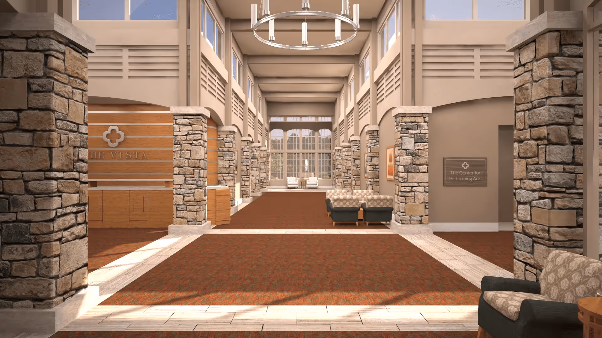 A spacious and well-lit interior hallway with stone pillars and large windows. The hallway features a red carpet with a beige border, comfortable armchairs with patterned upholstery, and a chandelier hanging from the ceiling. Signs indicate areas named 'The Vista' and 'The Center for Performing Arts'.