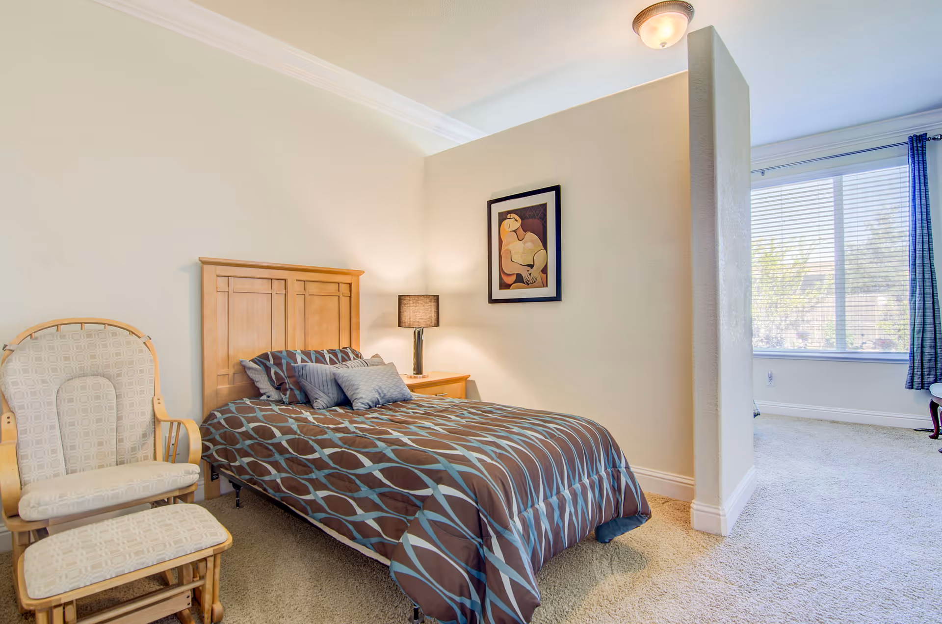 A bedroom with a wooden headboard bed covered in a brown and blue patterned comforter and several pillows. Next to the bed is a wooden nightstand with a lamp and a framed abstract painting on the wall above it. To the left of the bed is a cushioned rocking chair with a matching footrest. The room has beige walls, carpeted floor, and a partial wall separating the bed area from a window with blue curtains letting in natural light.