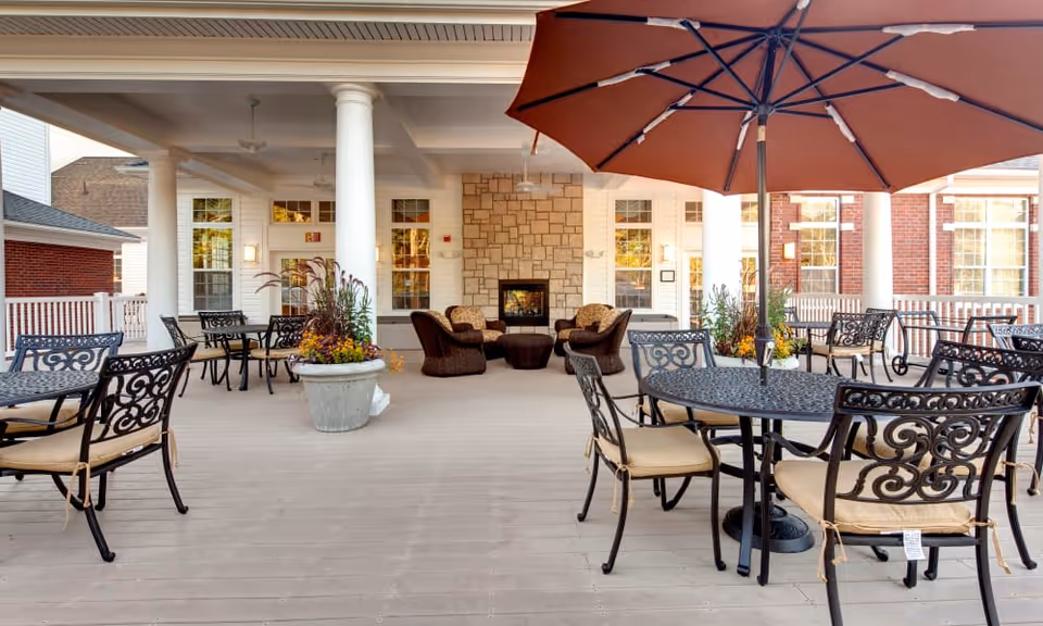 Outdoor covered patio area with multiple black metal tables and chairs with beige cushions, a large brown umbrella over one table, potted plants with flowers, and a stone fireplace with seating in the background. The area is part of a building with white columns and large windows.