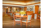 A warm and inviting dining area in a senior living facility with wooden tables and chairs. A round table in the foreground is decorated with a vase of yellow flowers. The room features soft lighting, wooden cabinetry, and a stone accent wall with a clock and decorative items.