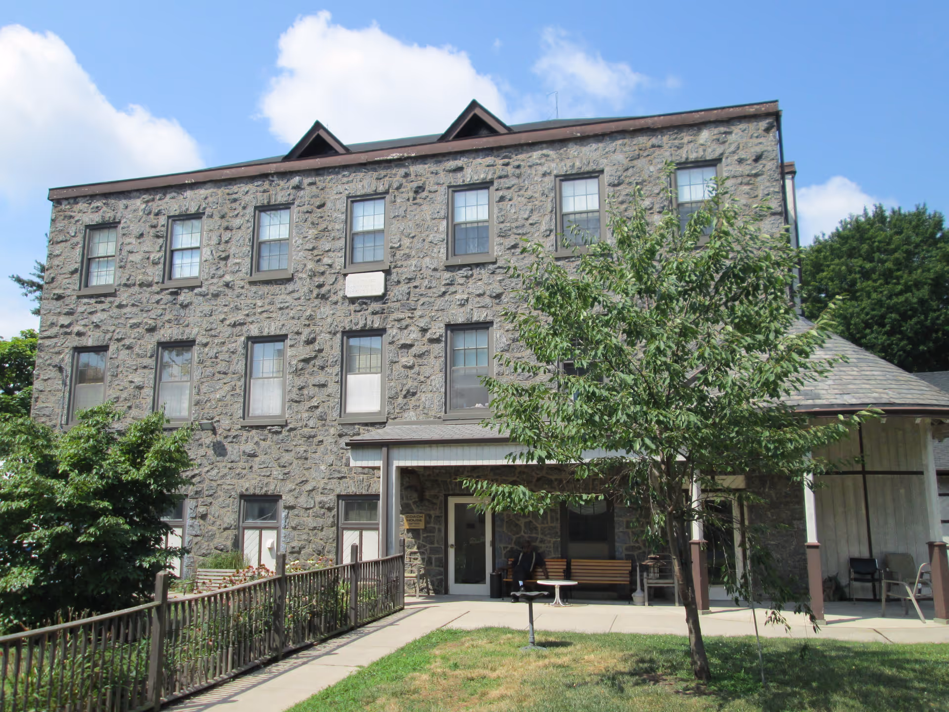 Exterior view of a three-story stone building with multiple windows and a covered entrance. There is a small tree and some greenery in front of the building, along with a paved walkway and a bench near the entrance. The sky is partly cloudy.