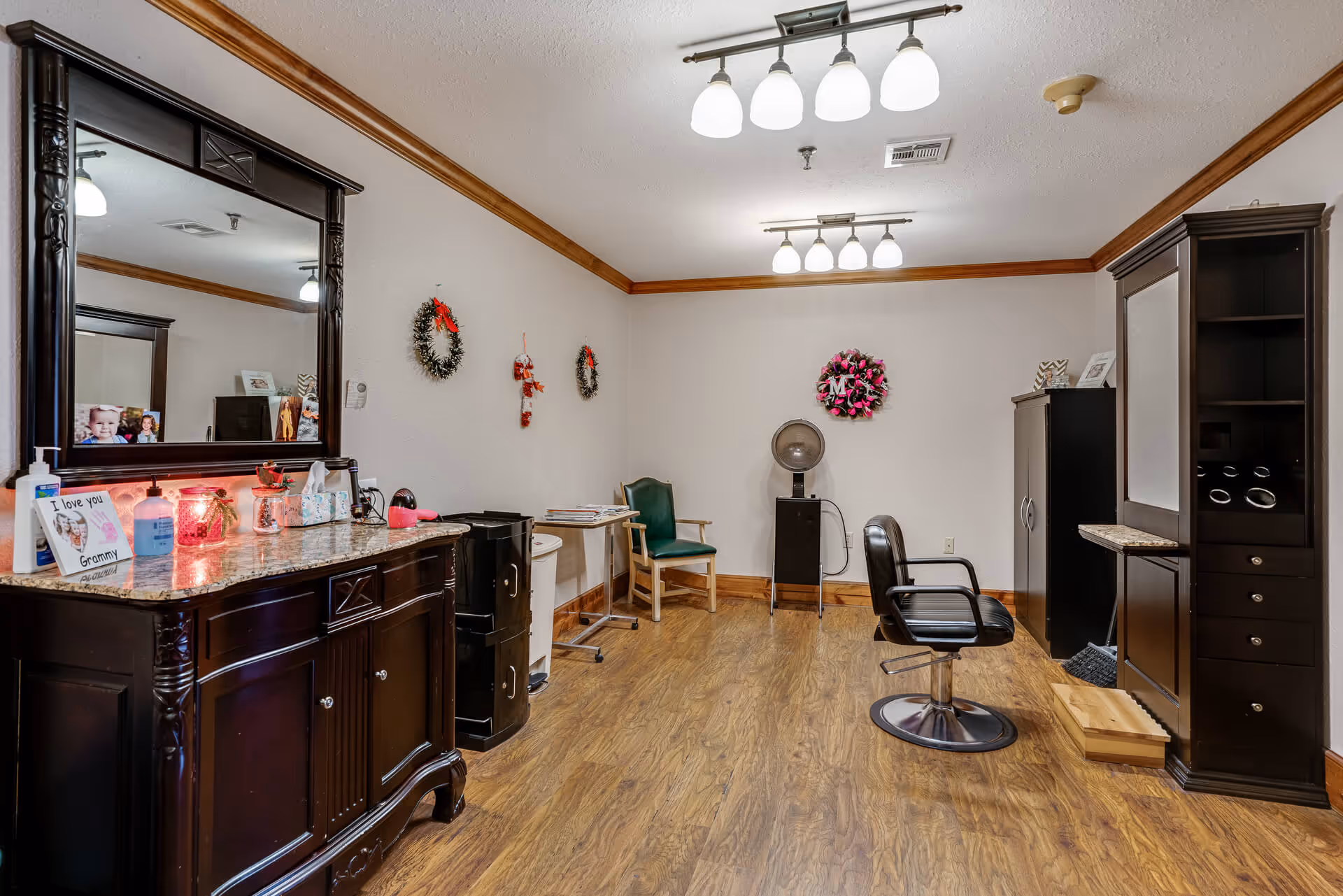 A bright senior living salon room with wood floors, a styling chair, mirrors, cabinets, and wreath decorations on the walls.