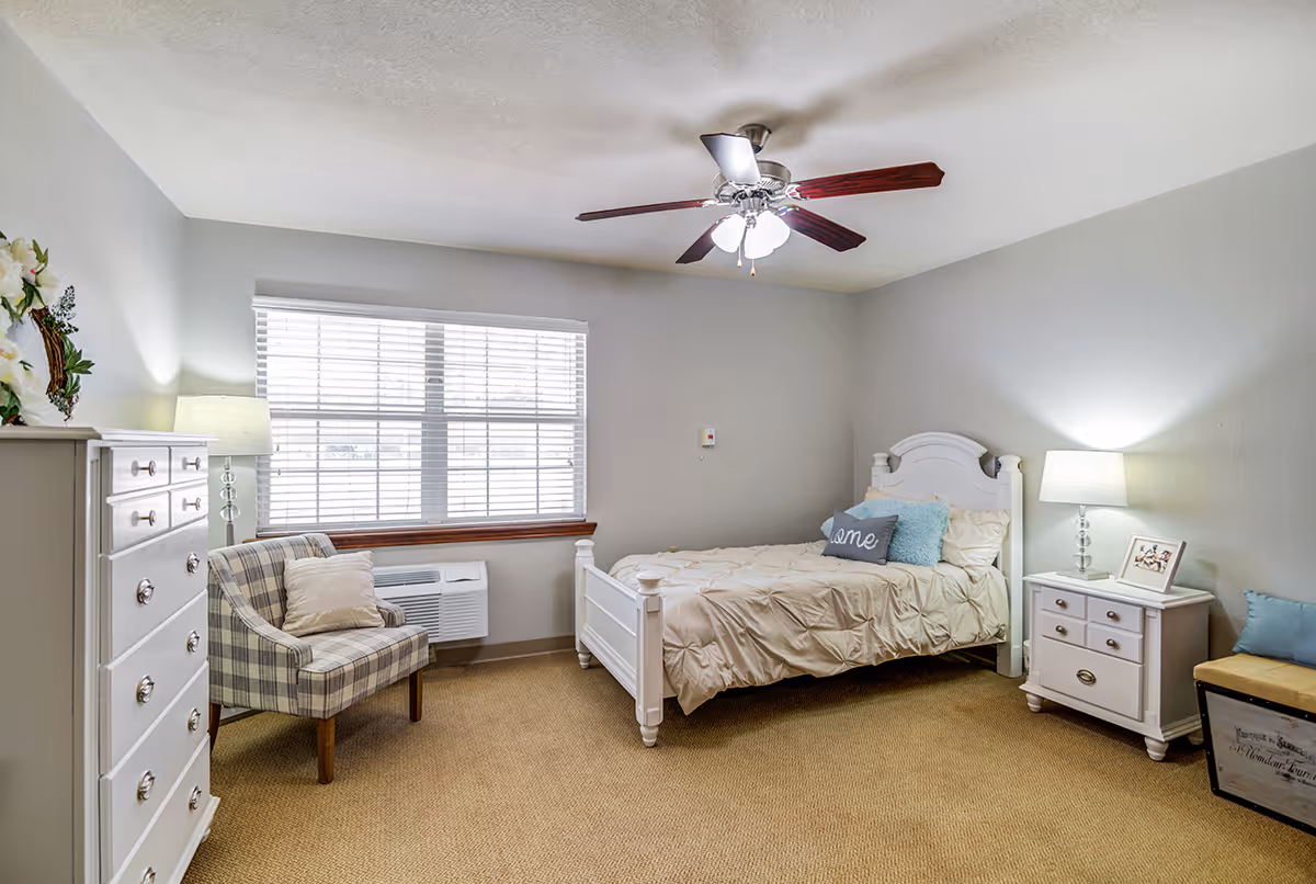 A cozy bedroom with a white wooden bed featuring beige bedding and decorative pillows. There is a white nightstand with a lamp and a framed photo on the right side of the bed. On the left side of the room, there is a white dresser with a floral arrangement on top and a plaid upholstered armchair with a cushion. A large window with white blinds lets in natural light, and a ceiling fan with lights is mounted on the ceiling. The room has beige carpet and light gray walls.