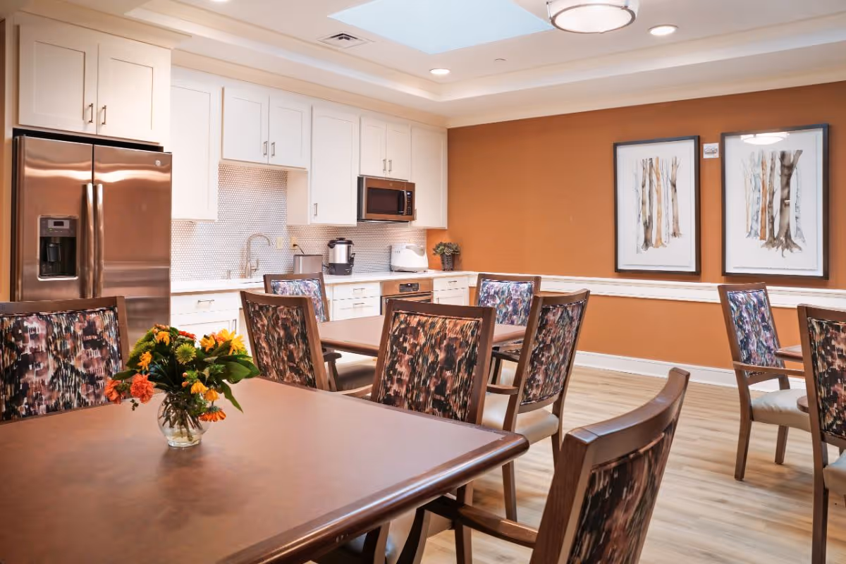 A dining area in a senior living facility featuring several tables with floral centerpieces and chairs with patterned upholstery. The room has a warm orange accent wall with two framed tree artwork pieces. The background shows a kitchen area with white cabinets, a stainless steel refrigerator, a microwave, and various kitchen appliances. The space is well-lit with ceiling lights and a skylight.
