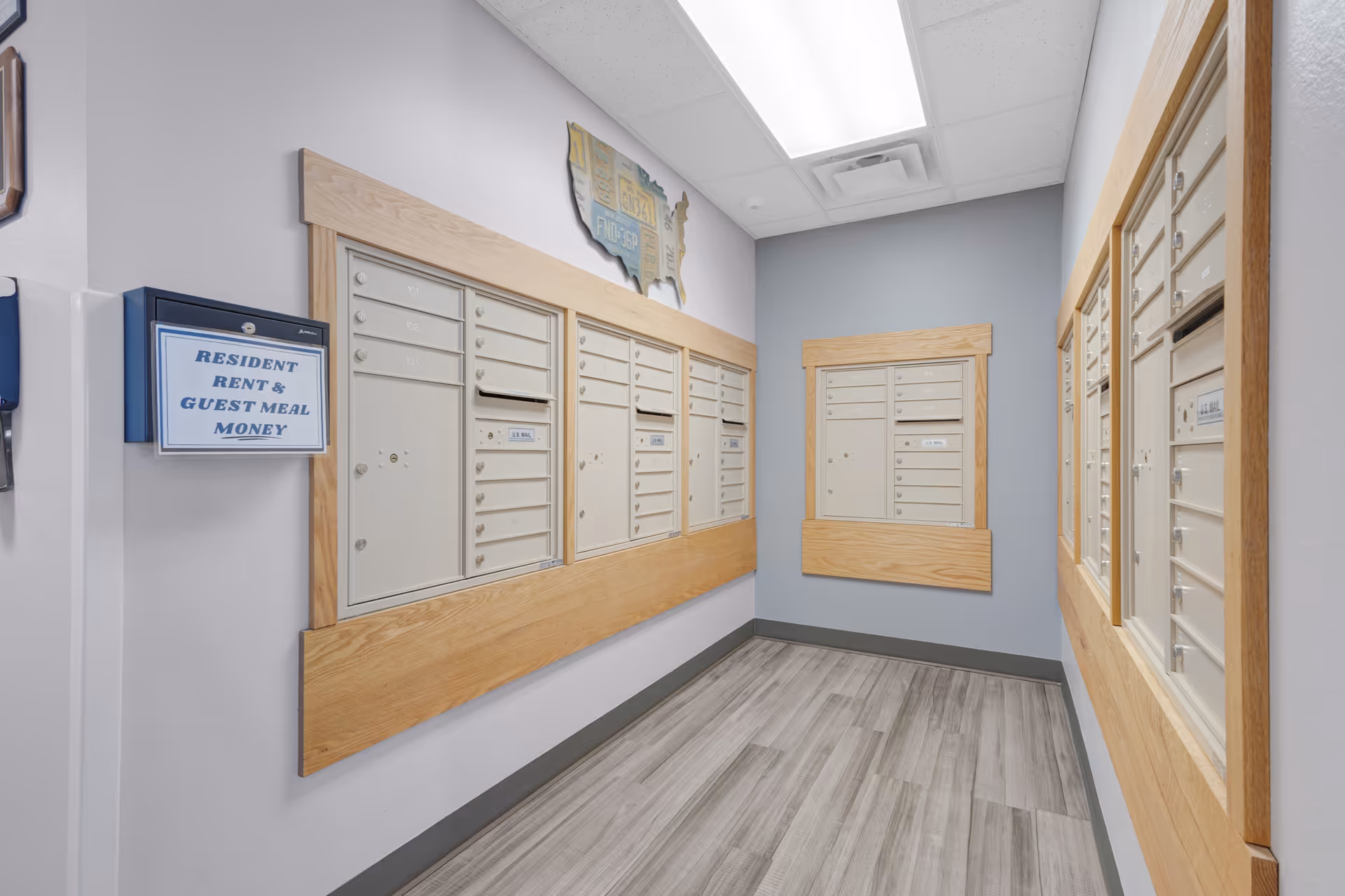 A hallway with multiple beige mailboxes mounted on light purple and gray walls, framed with light wood trim. A sign on the left wall reads 'Resident Rent & Guest Meal Money.' The floor has gray wood-like tiles, and the ceiling has white panels with fluorescent lighting.