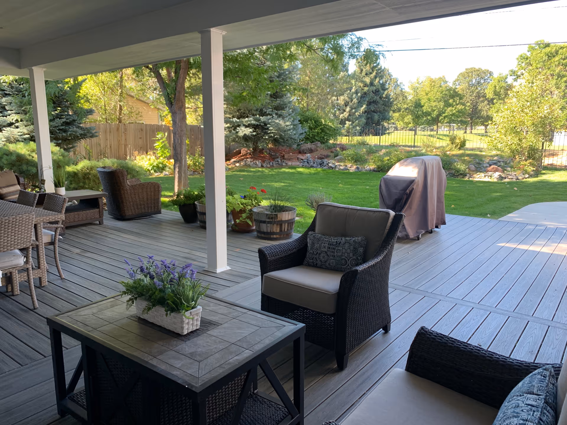 Covered outdoor patio area with wicker chairs and a table with a flower arrangement, overlooking a green lawn with trees and a covered grill.