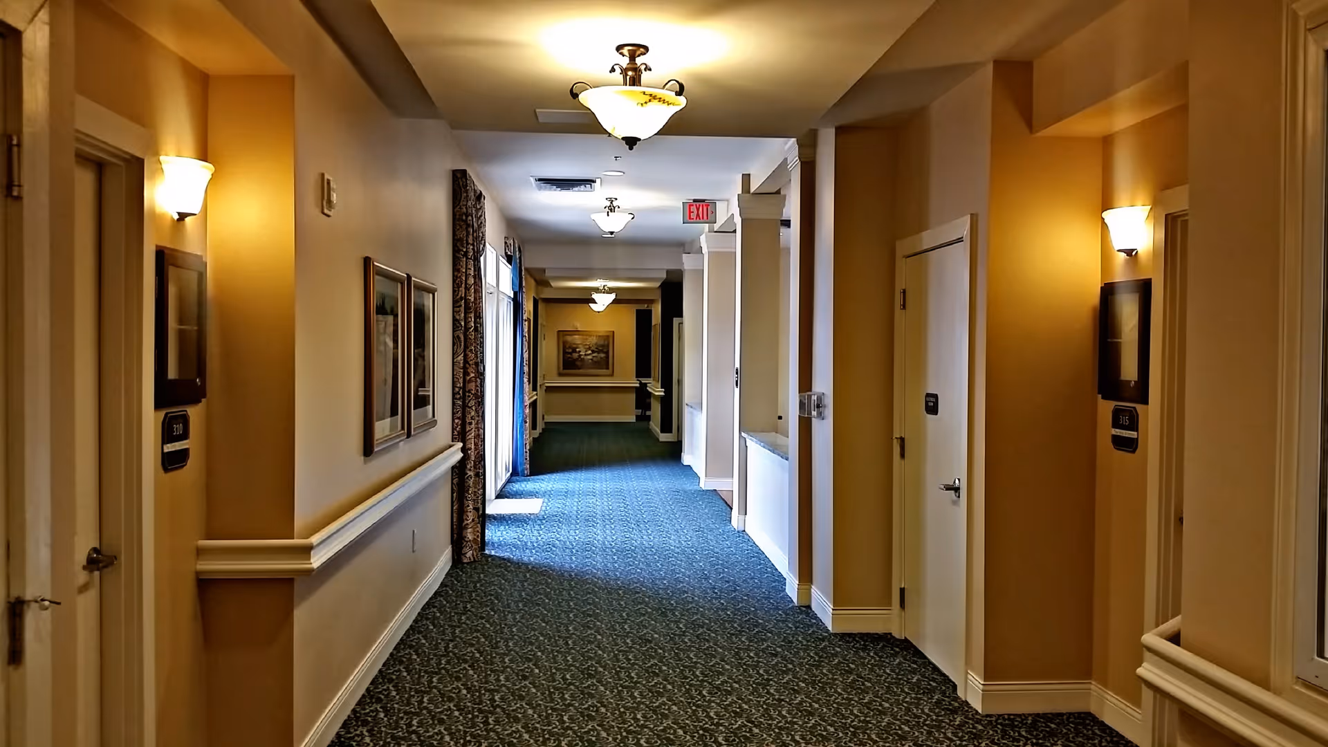 A well-lit hallway in a senior living facility with beige walls, patterned carpet, wall sconces, framed pictures, and several doors on both sides. There is an exit sign visible at the far end of the corridor.