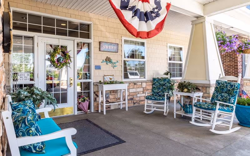 Covered front porch of a senior living facility with rocking chairs, potted plants, decorative bunting and glass entrance doors.