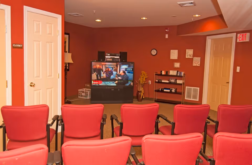 A small media room with red walls and red chairs arranged in rows facing a television. The TV is on and shows a program with people. There is a bookshelf with books and a vase with yellow flowers next to the TV. Two doors are visible, one labeled 'Closet' and the other near an exit sign.