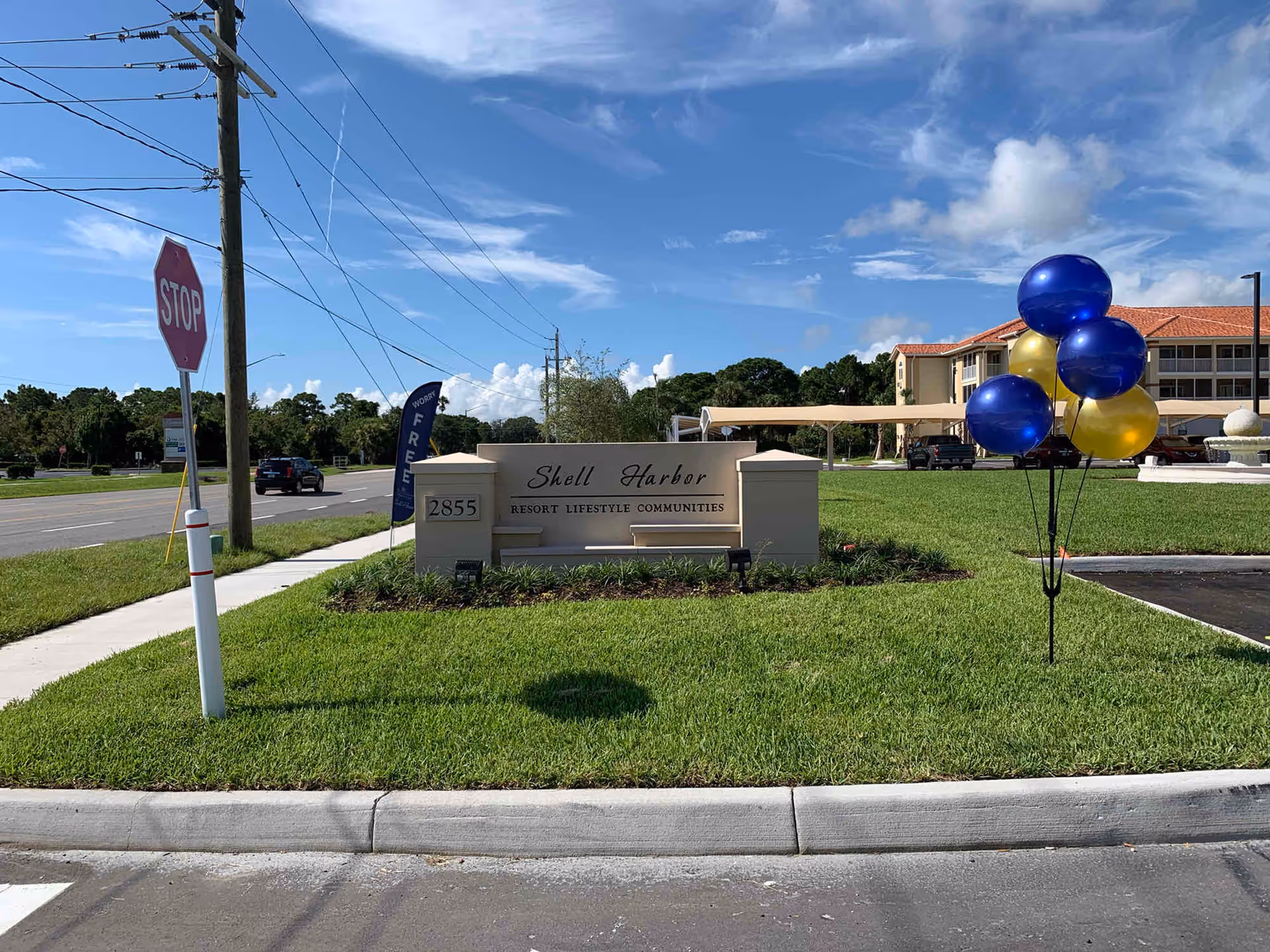 Outdoor view of the entrance sign for Shell Harbor Resort Lifestyle Communities with the address 2855. The sign is set on a grassy area next to a road with a stop sign and utility poles. Blue and gold balloons are tied to a stand on the grass near the sign. In the background, there is a multi-story building with a red roof and a covered parking area.