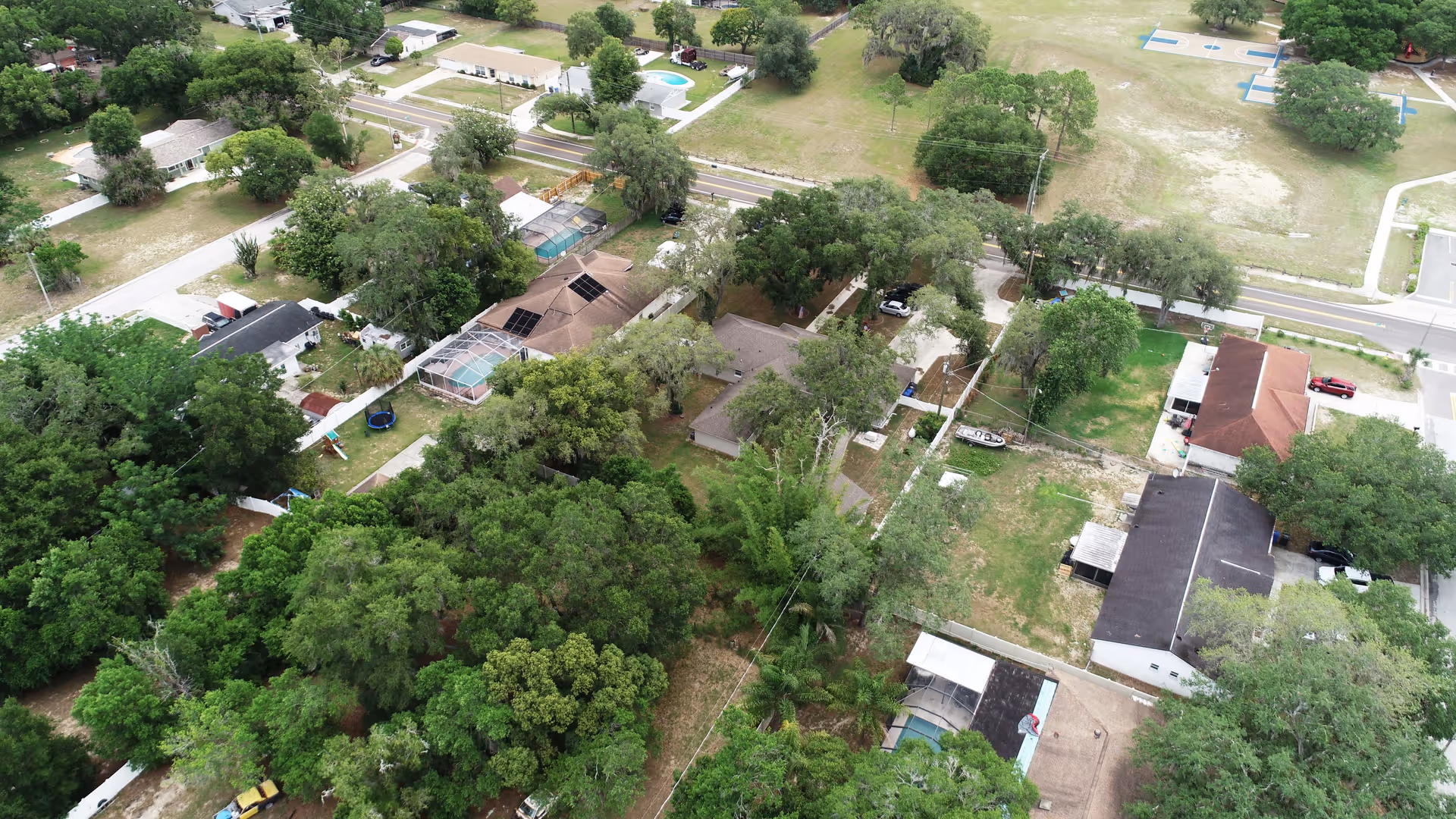 Aerial view of a residential neighborhood with several houses, trees, lawns, and a road running through the area. Some houses have swimming pools and fenced yards. There is a basketball court visible in the upper right corner.