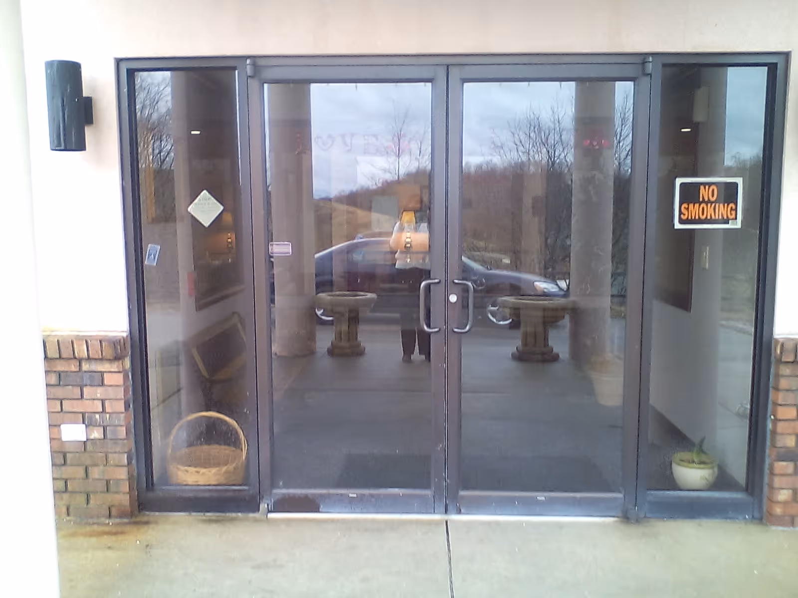 Glass double doors entrance to a building with brick pillars on either side. There is a 'NO SMOKING' sign on the right glass panel and a basket on the floor inside the left glass panel. A car and some trees are reflected in the glass doors.