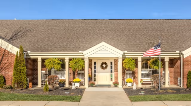 Front exterior view of a single-story brick building with a peaked roof, white columns, and a covered porch. The entrance door is centered with a wreath, flanked by windows with hanging green plants. There are small shrubs and flowers planted along the front, and an American flag is displayed on a flagpole to the right of the entrance.