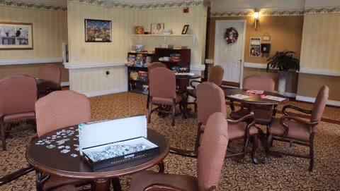 Community activity room with round tables and upholstered chairs, a jigsaw puzzle on a table and shelves of board games against the wall.